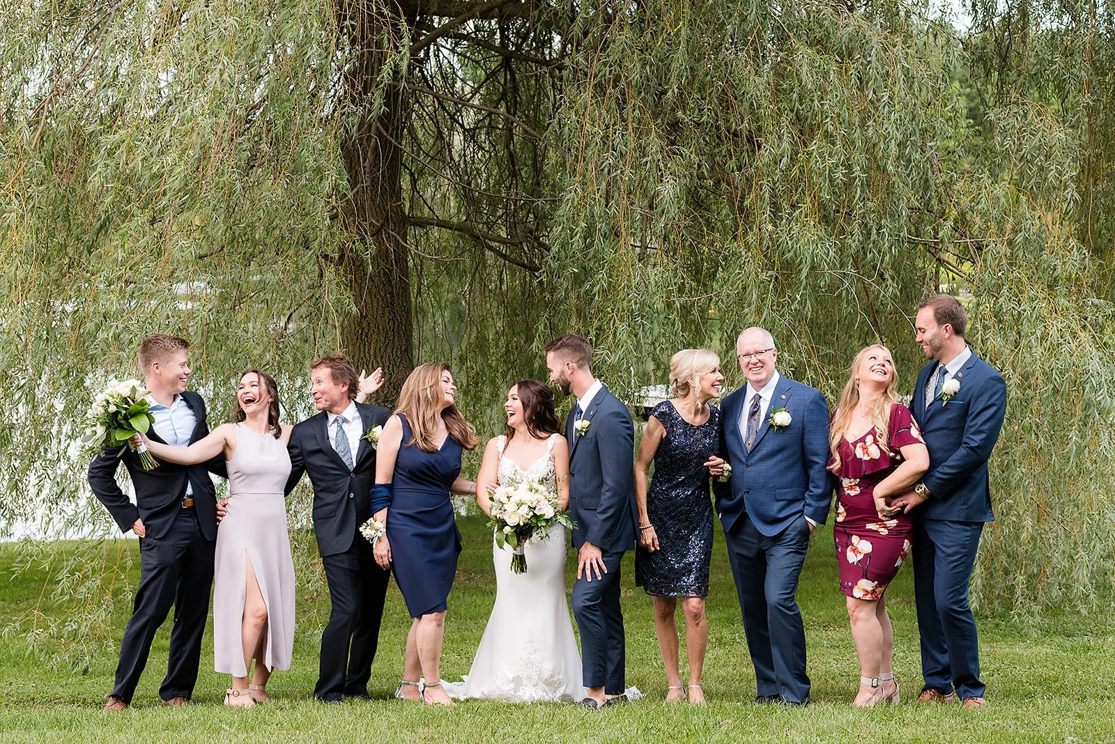 A group of people dressed in formal attire standing outdoors near a large tree, smiling, and sharing a joyful moment at a wedding celebration.