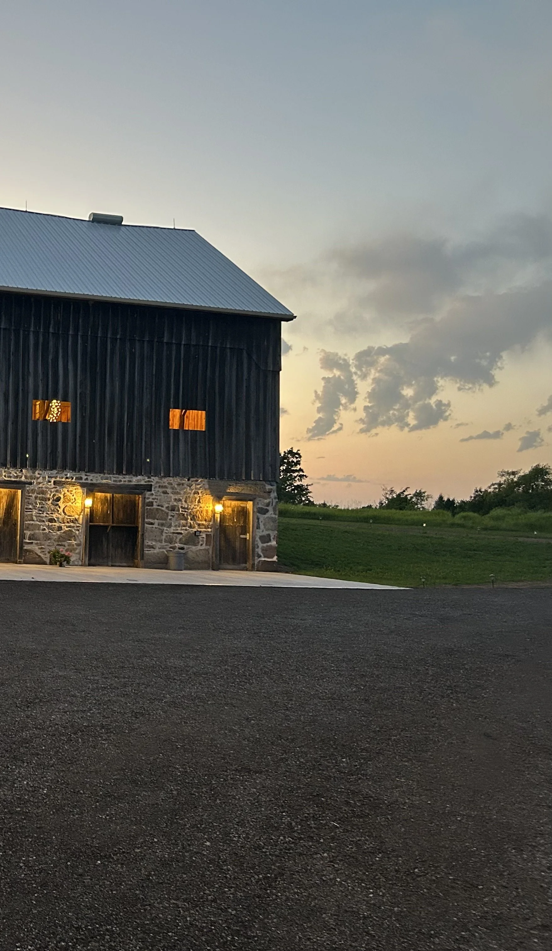 A black barn with stone foundation, illuminated by outdoor lights at dusk, green grass and cloudy sky in the background.