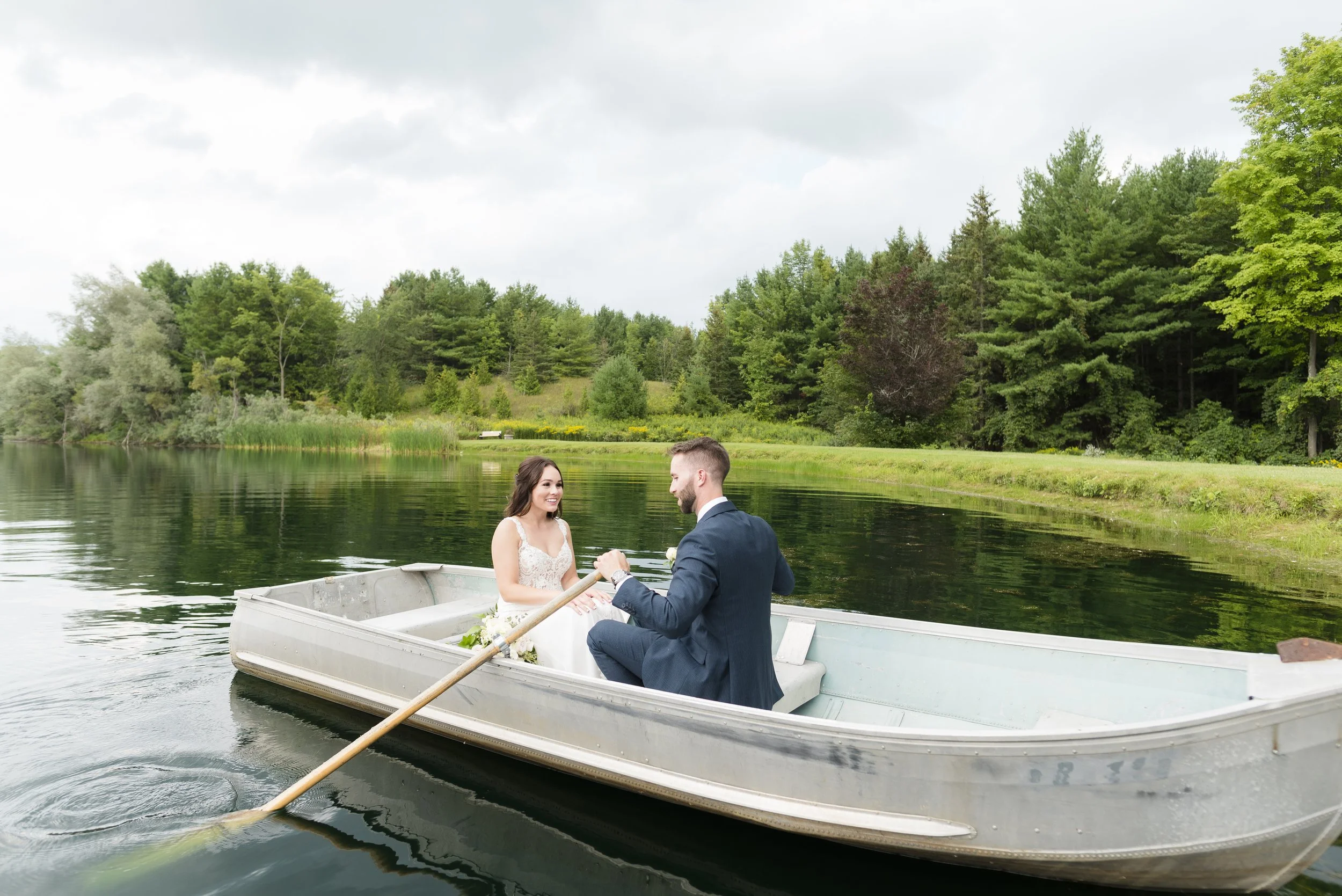 A bride and groom on a small boat on a lake, surrounded by green trees.