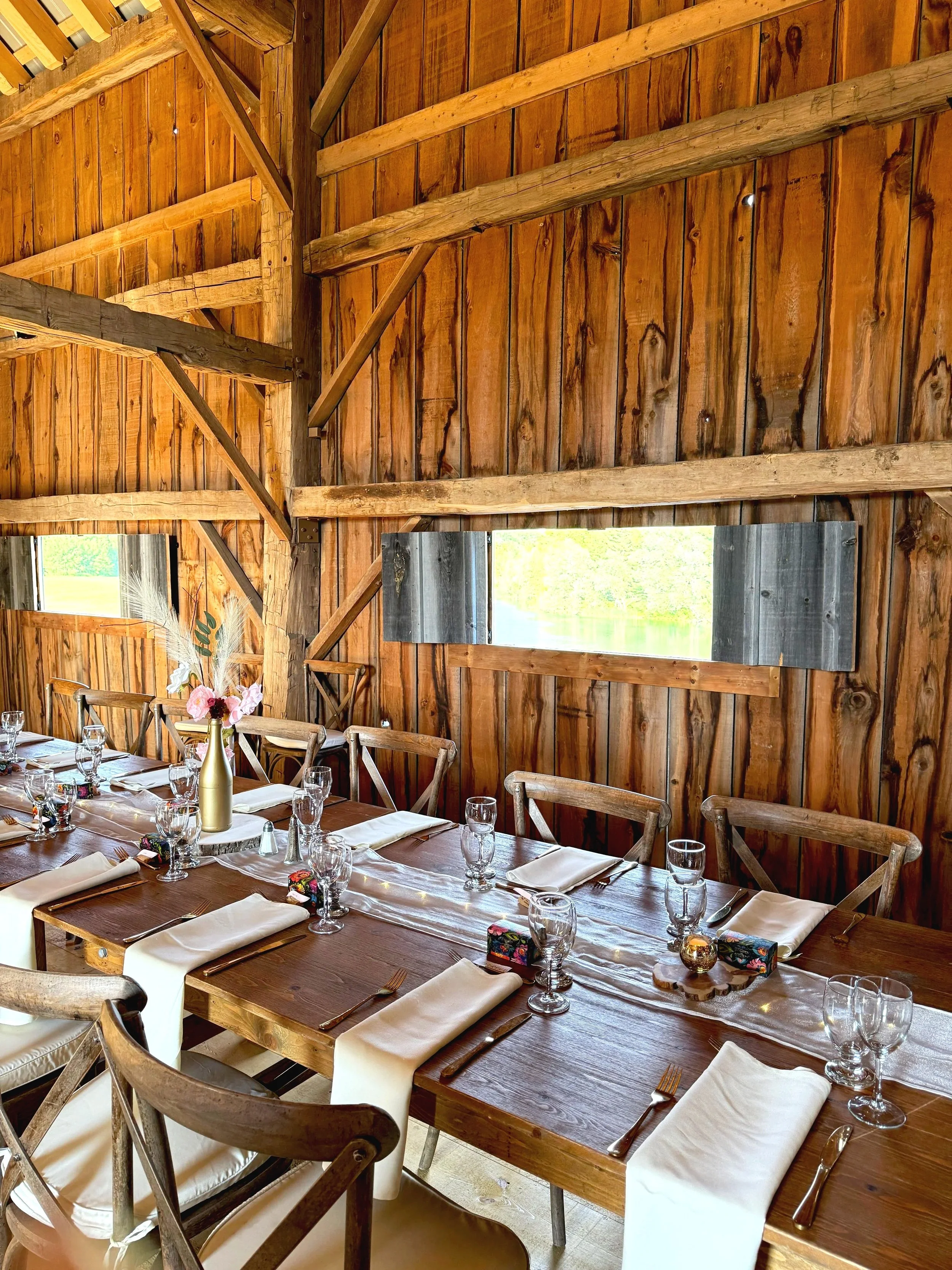 A rustic wooden dining room with a long table set for a meal, decorated with a centerpiece, glasses, napkins, and silverware.