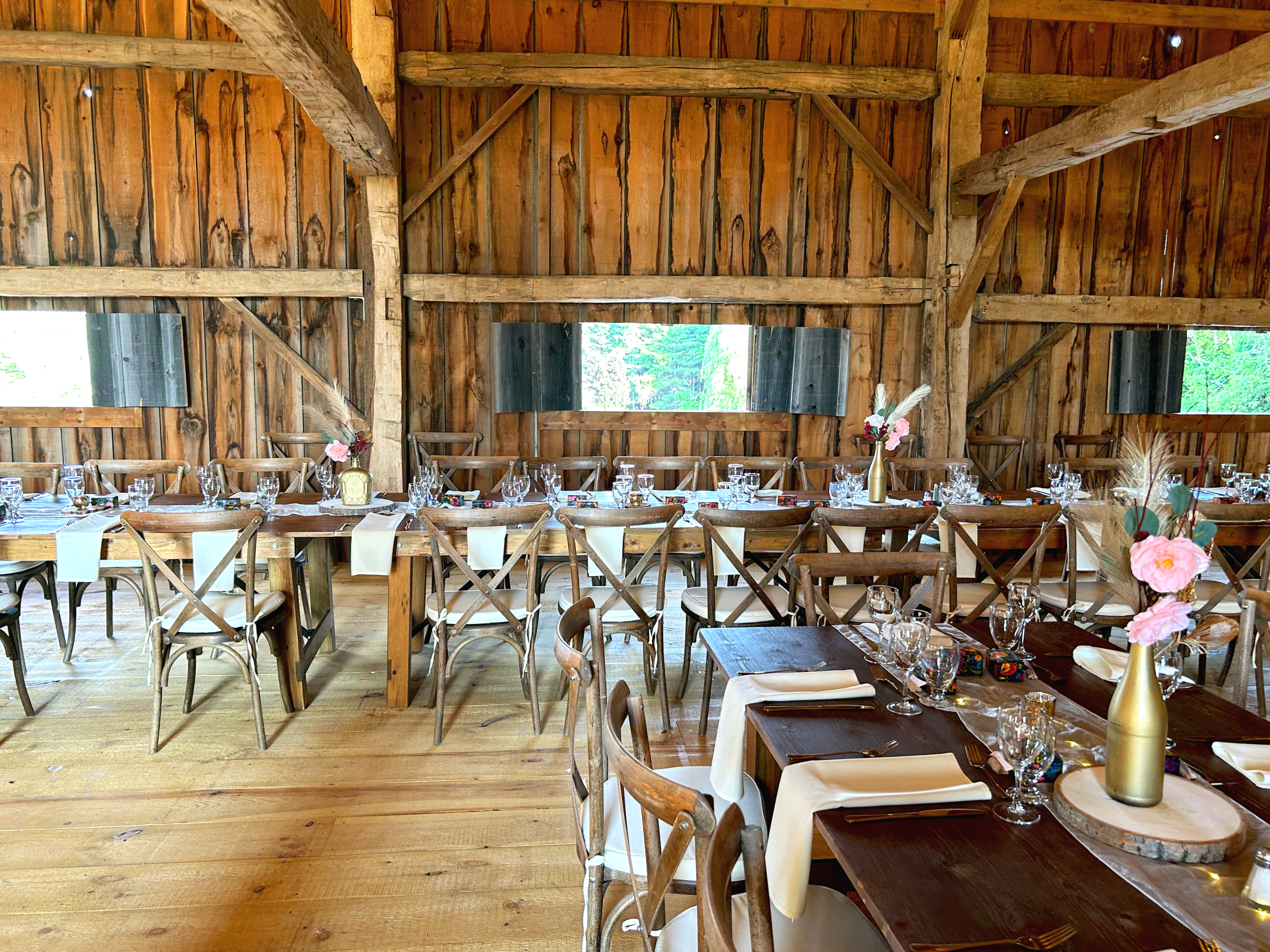 Inside a rustic wooden barn decorated for a wedding reception with long tables set with plates, glasses, and napkins, and floral centerpieces in gold vases.