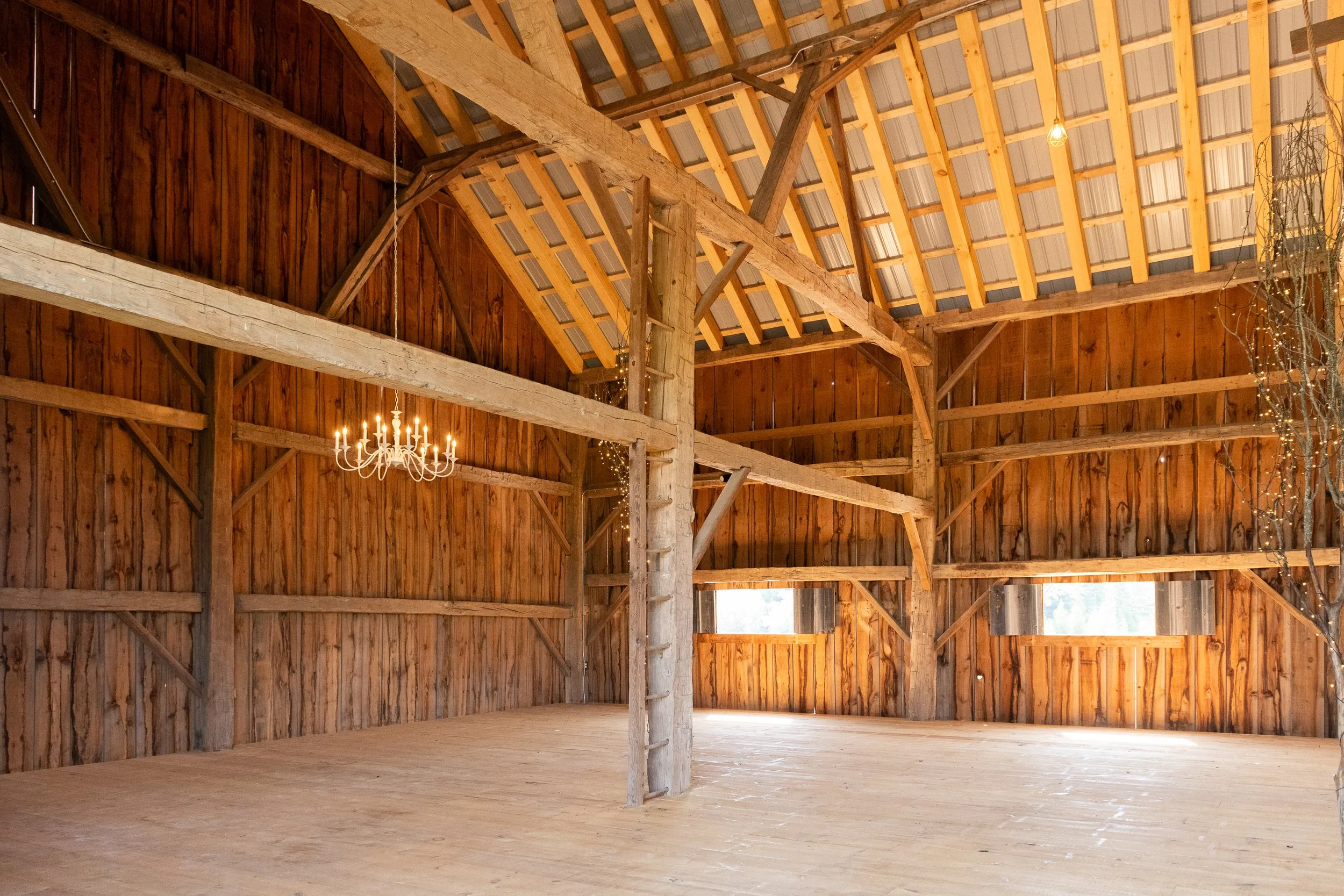 Empty wooden barn interior with high ceiling, exposed beams, a chandelier, string lights, and small windows.