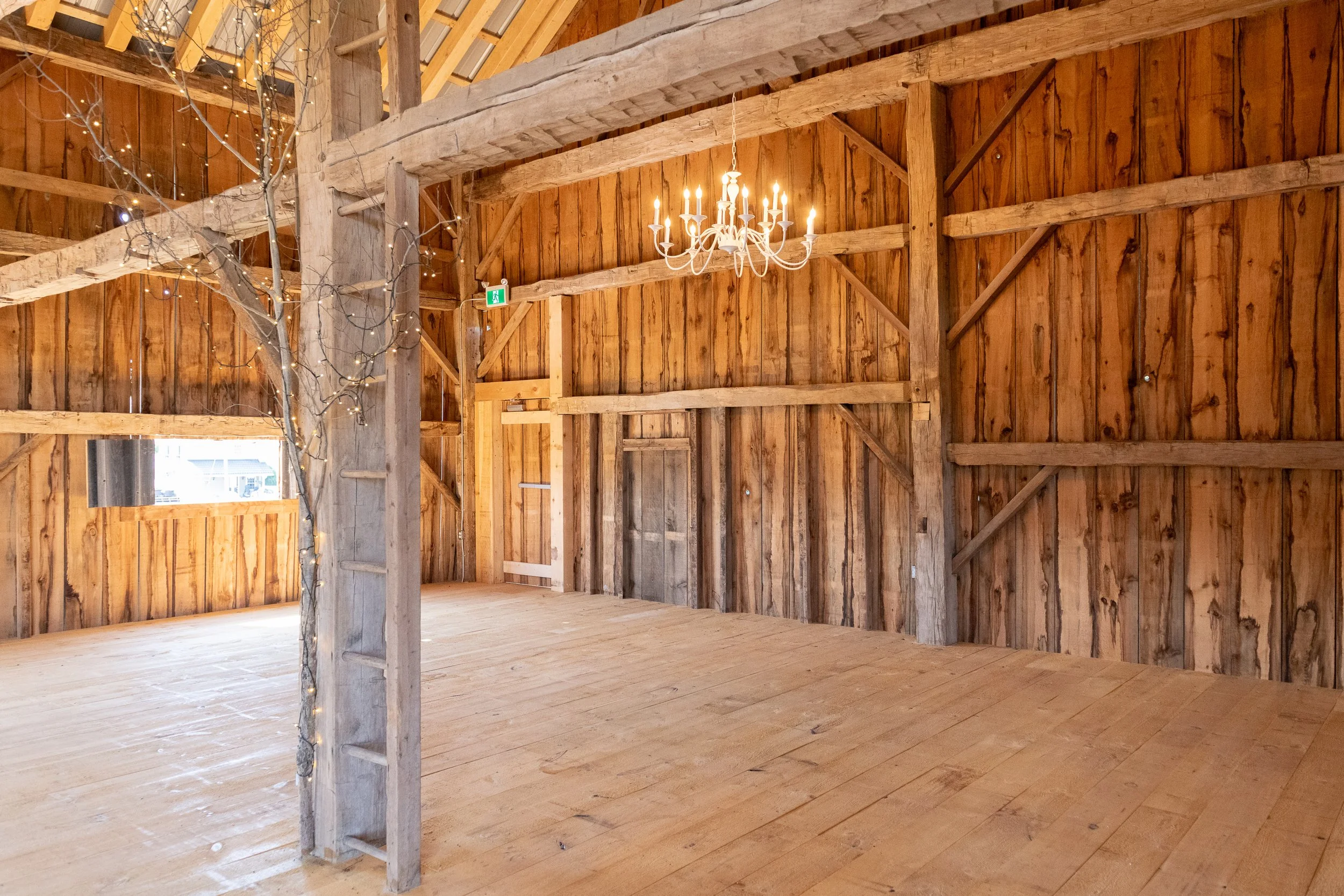 Interior of a rustic wooden barn with a chandelier hanging from the ceiling, string lights wrapped around a wooden post, and a small window on the left side.