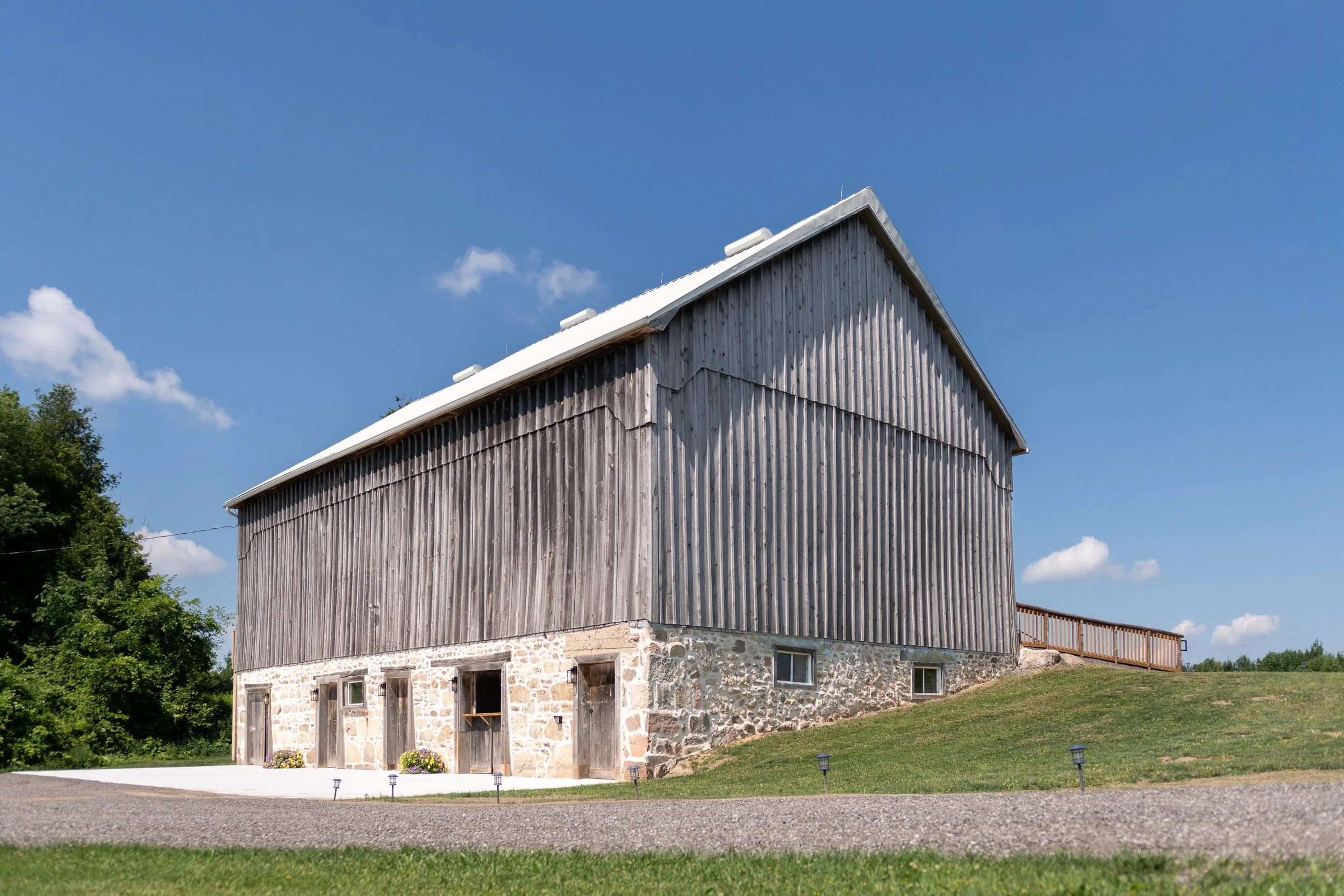 A rustic barn with stone foundation, wooden upper structure, and metal roof on a grassy lawn under a blue sky with a few clouds.