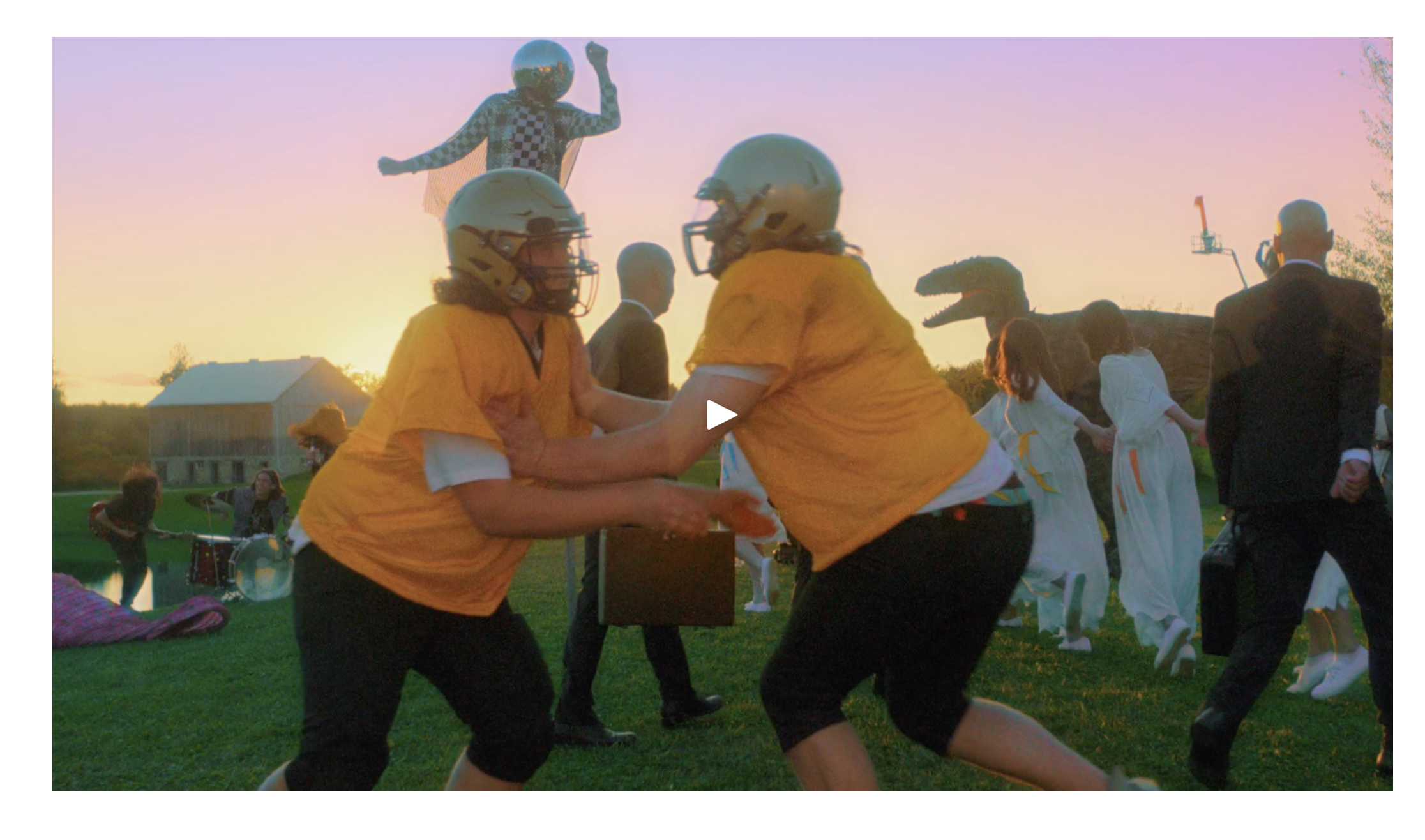 Two people in yellow shirts and helmets playfully pushing each other at an outdoor party during sunset, with children holding hands in the background and a person standing near a sound system, a barn, and a juggling clown on a unicycle in the distanc