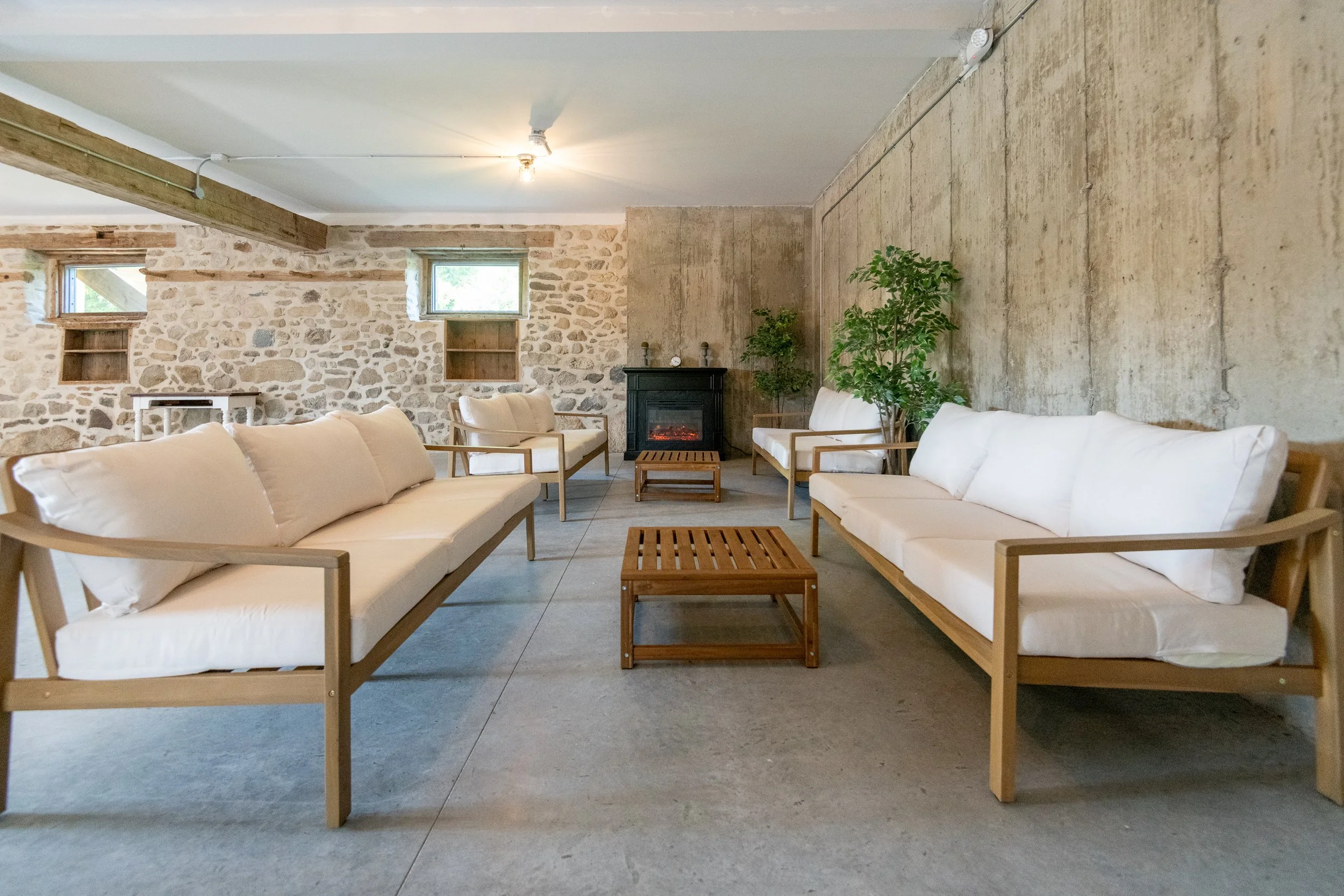 A cozy living room with four white cushioned sofas with wooden frames arranged around a small wooden coffee table, in front of a black fireplace on a beige wall. The room has a stone wall with small windows, two green potted plants, and a concrete fl