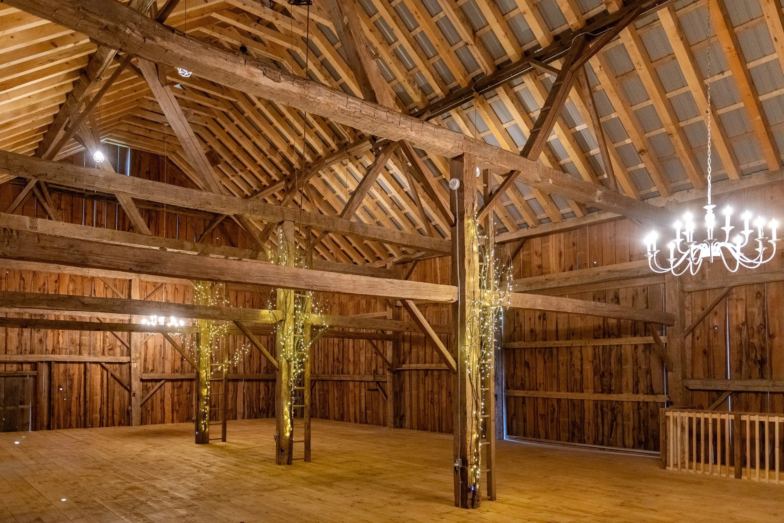 Interior of a large wooden barn with Christmas lights wrapped around the support beams and a chandelier hanging from the ceiling.