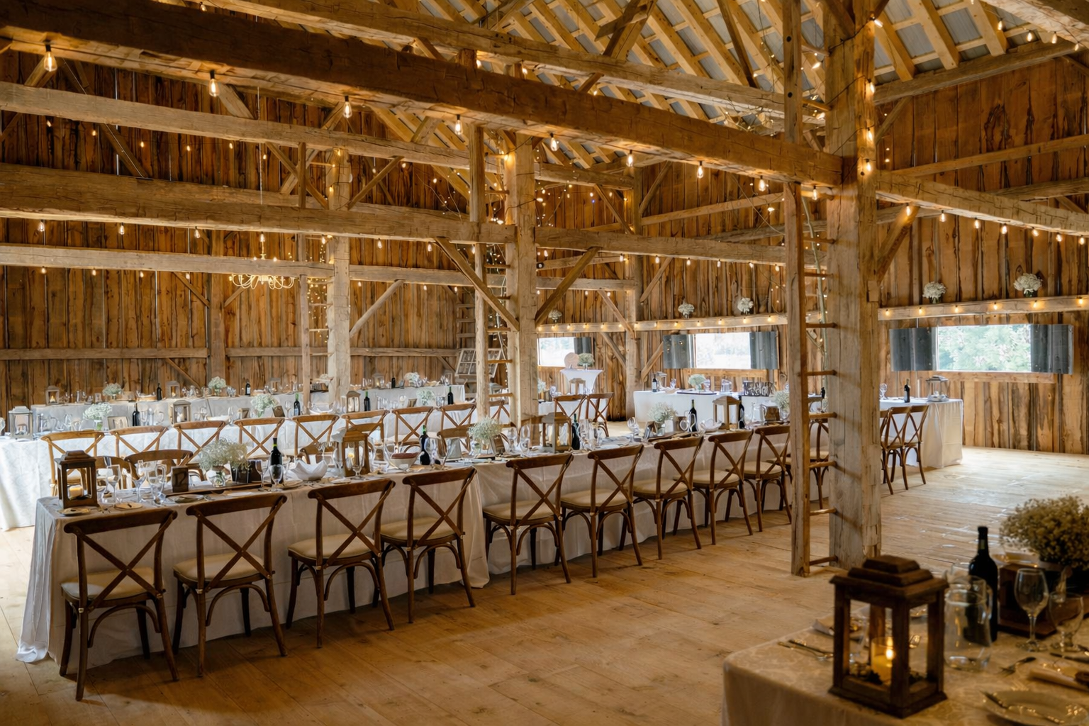 Elegant barn wedding reception at The Hummingbird in Erin, Ontario featuring long harvest tables, wooden cross-back chairs, soft candlelight, and warm string lighting beneath restored heritage beams.