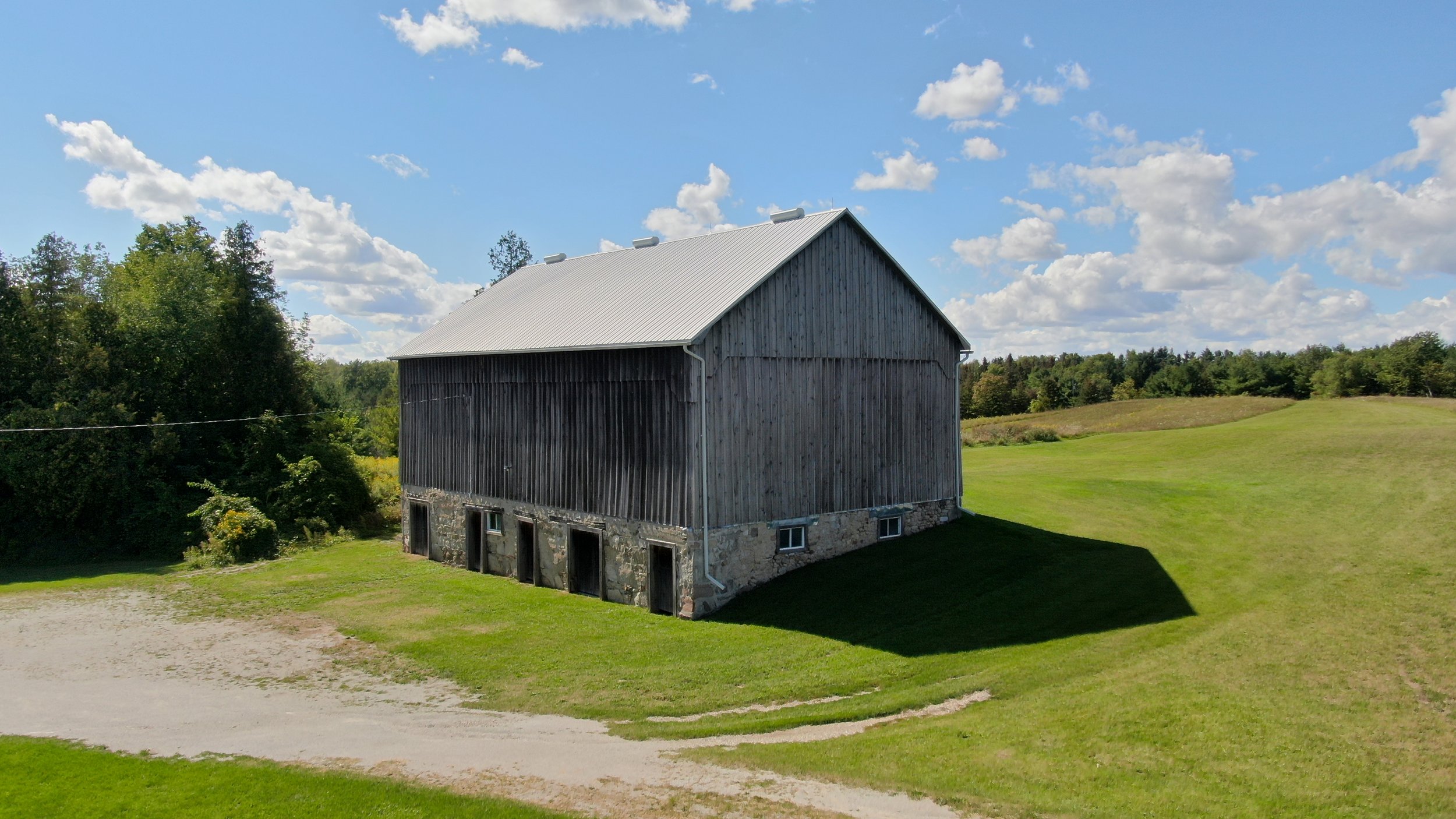 A large wooden barn with a metal roof sits on a grassy field under a partly cloudy sky.