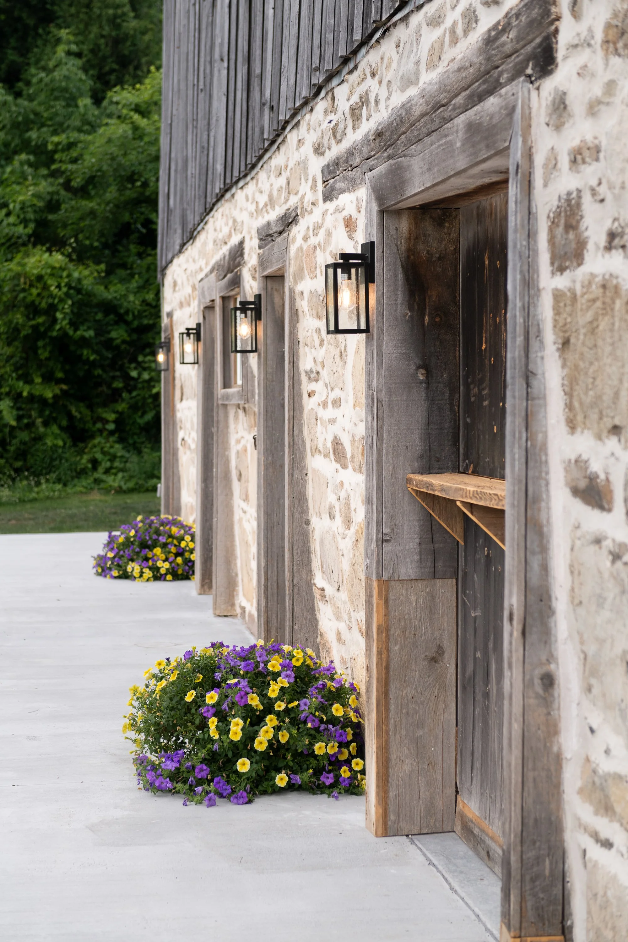 Stone and wood exterior wall of a rustic building with black wall-mounted lanterns and colorful potted flowers on the ground.