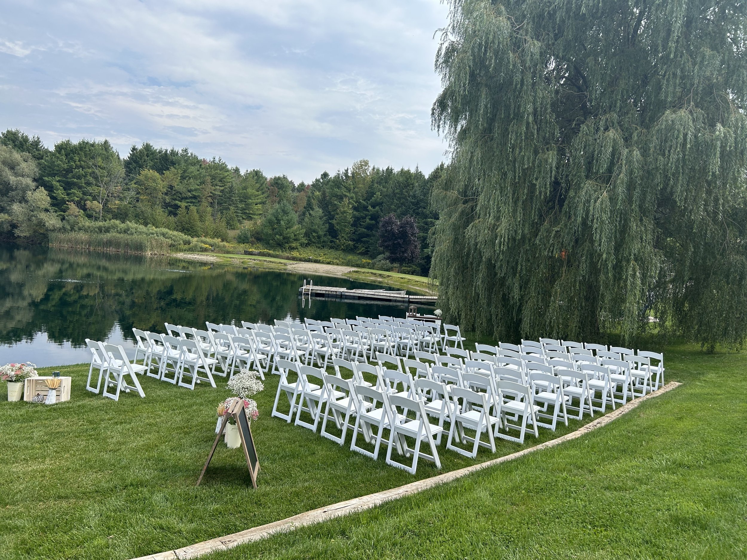 A romantic lakeside wedding ceremony at The Hummingbird in Erin, Ontario, set beneath a graceful willow tree beside the pond. White ceremony chairs and lush surroundings create an intimate, nature-filled setting for an unforgettable outdoor celebrati