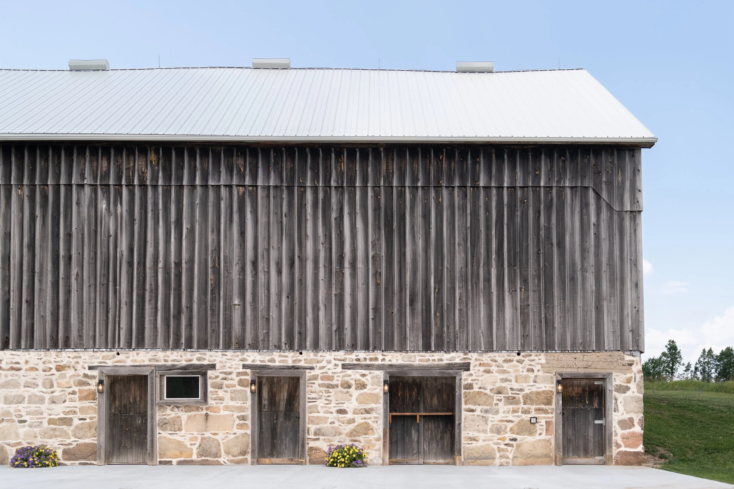 A rustic barn with a stone foundation, wooden doors, and a metal roof, set against a blue sky with grass on the right.