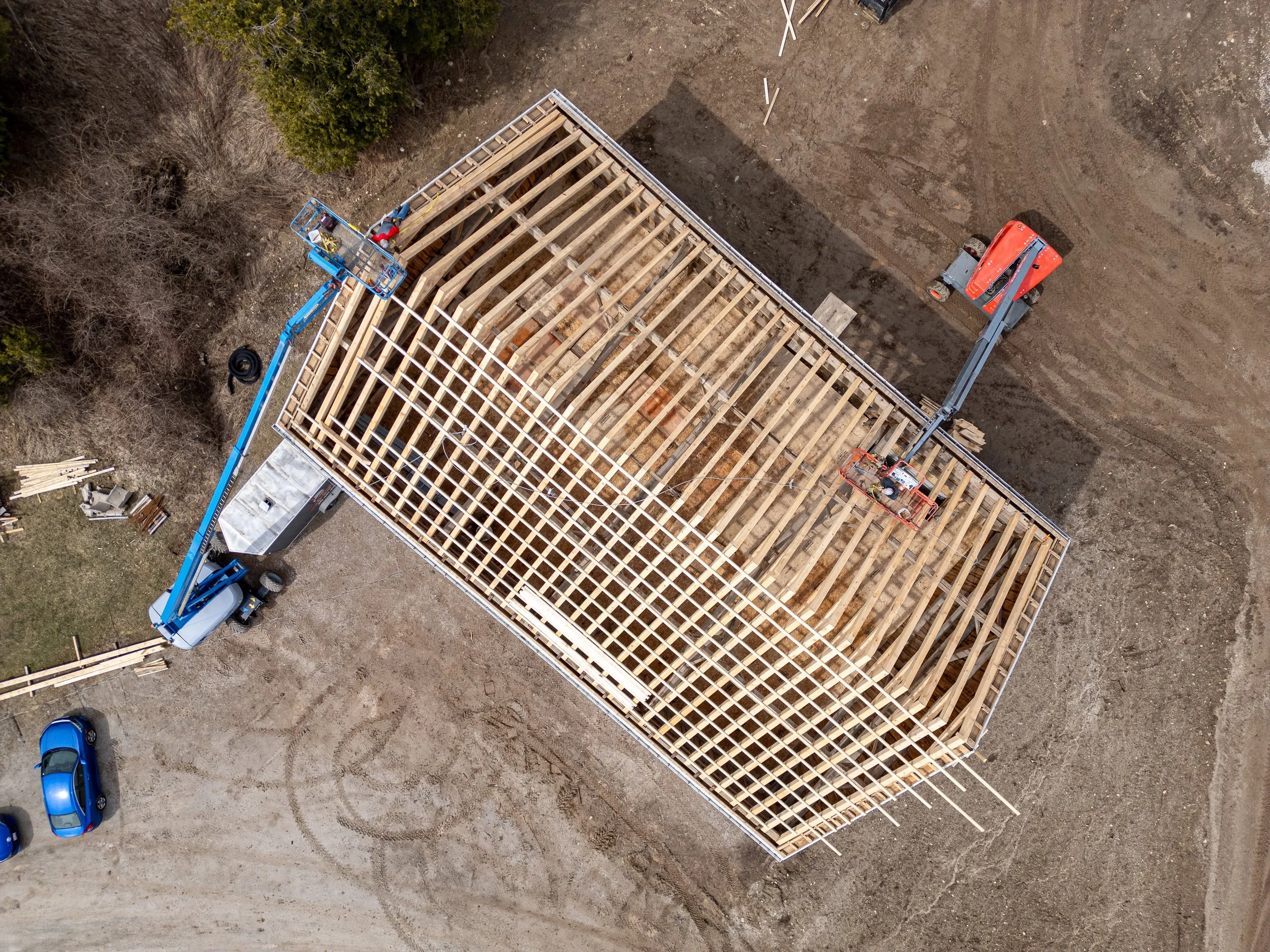 An aerial view of a building under construction with a wooden frame and roof trusses. Construction equipment, including a blue boom lift and an orange cherry picker, are present on-site along with a couple of parked blue cars and construction materia