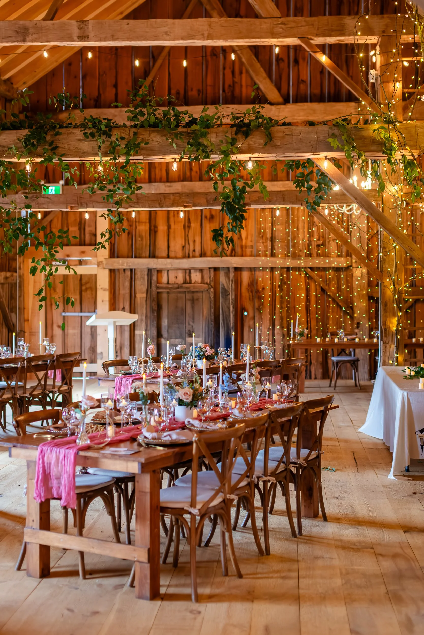 Styled barn wedding reception at The Hummingbird in Erin, Ontario, featuring harvest tables with blush table runners, cross-back chairs, soft candlelight, and lush greenery draped along exposed wooden beams. This romantic reception setup highlights t
