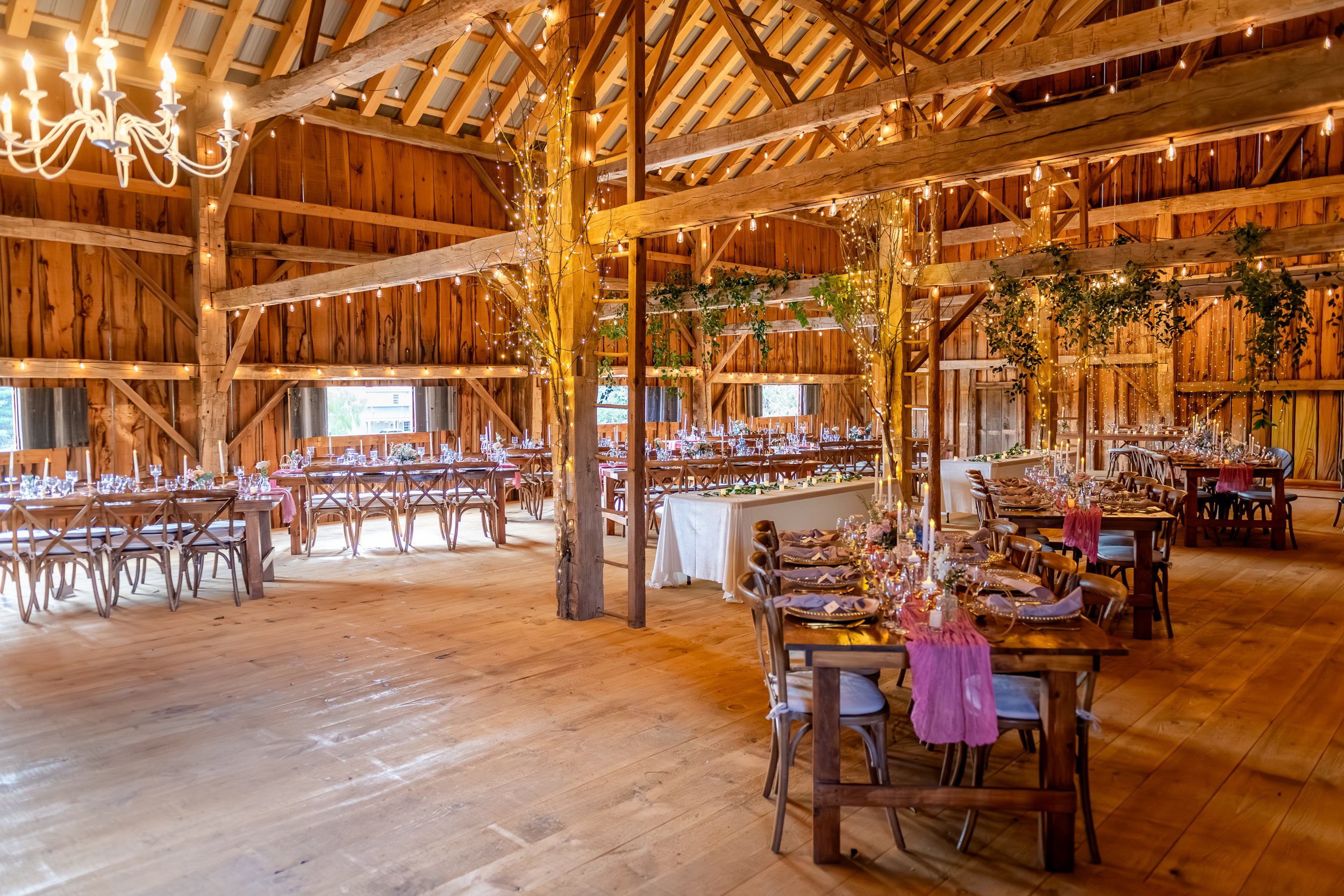 An elegant barn wedding reception at The Hummingbird in Erin, Ontario, featuring harvest tables, cross-back chairs, soft candlelight, blush table runners, and warm string lighting woven through exposed heritage beams. This romantic indoor wedding set