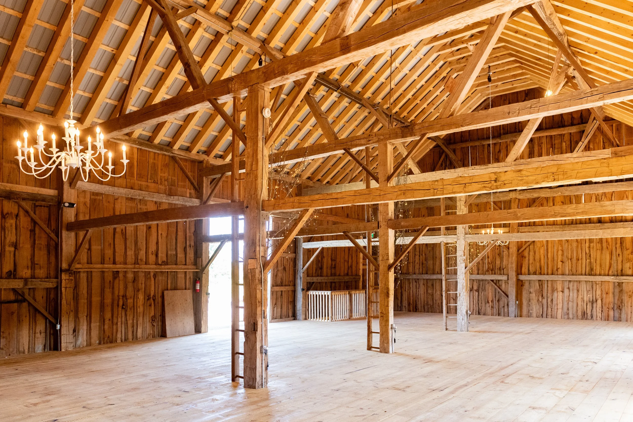 Interior of a large wooden barn with exposed beams, wood-paneled walls, and ceiling. Chandeliers hanging from the ceiling. Open door allowing sunlight in. Ladder against the wall. Decorated with string lights.