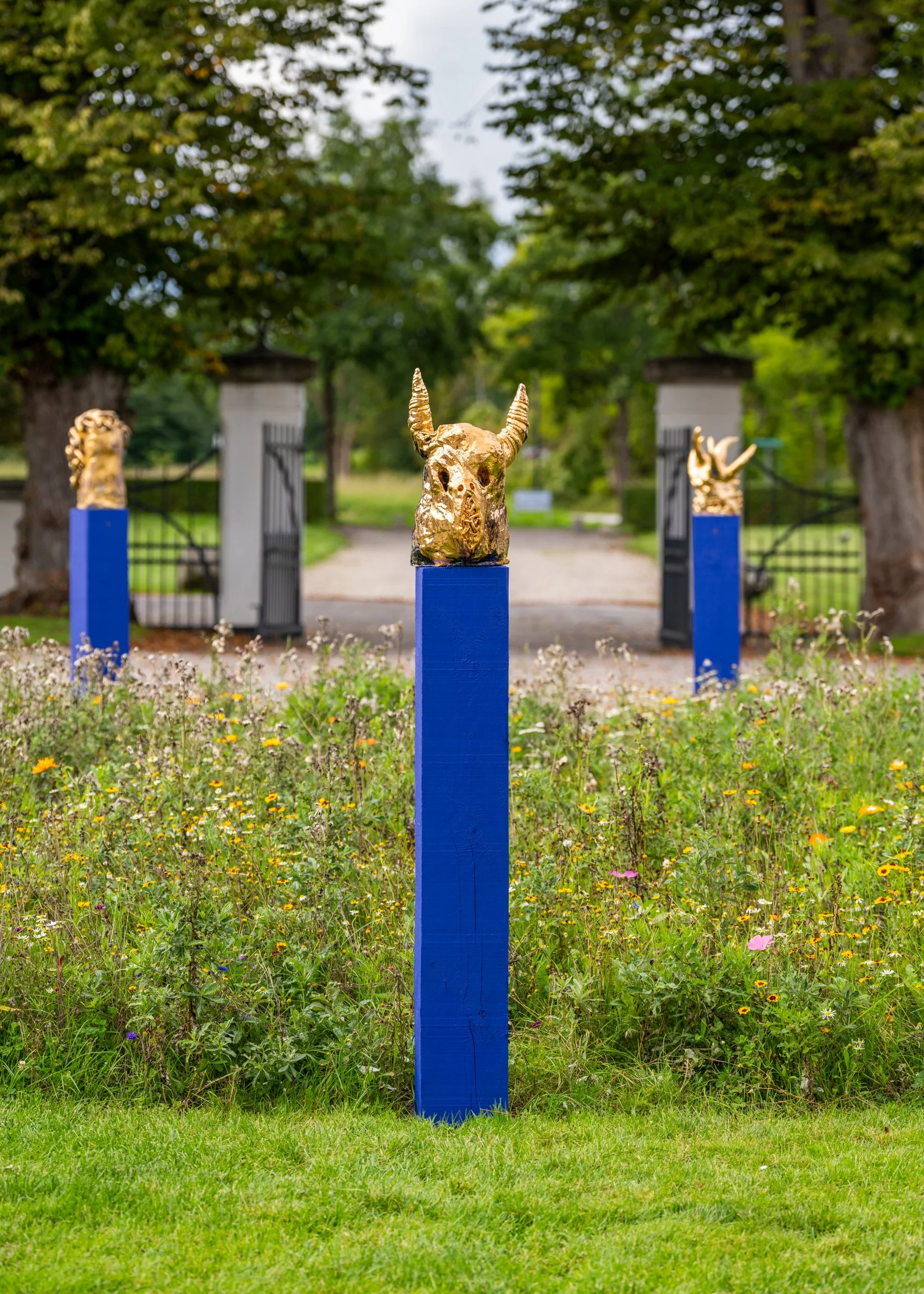 Three gold animal head sculptures on tall blue pedestals in a garden with green grass, wildflowers, and trees, with open gates in the background. Frederik Næblerød exhibition at Gl. Holtegaard, Denmark.