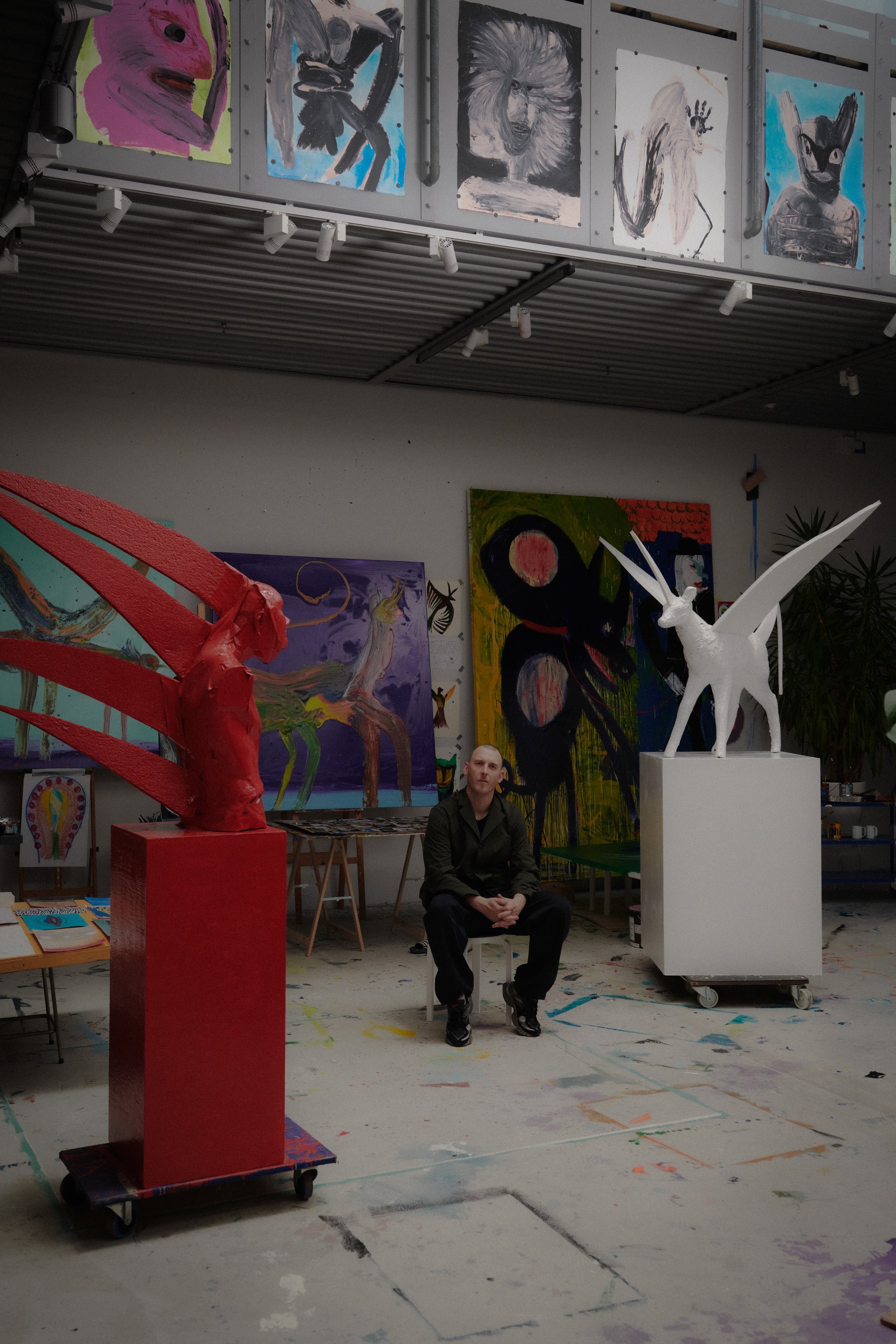 The Danish artist Frederik Næblerød sitting in an art studio surrounded by colorful and abstract sculptures and paintings, with artwork displayed on the walls and a paint-splattered floor.