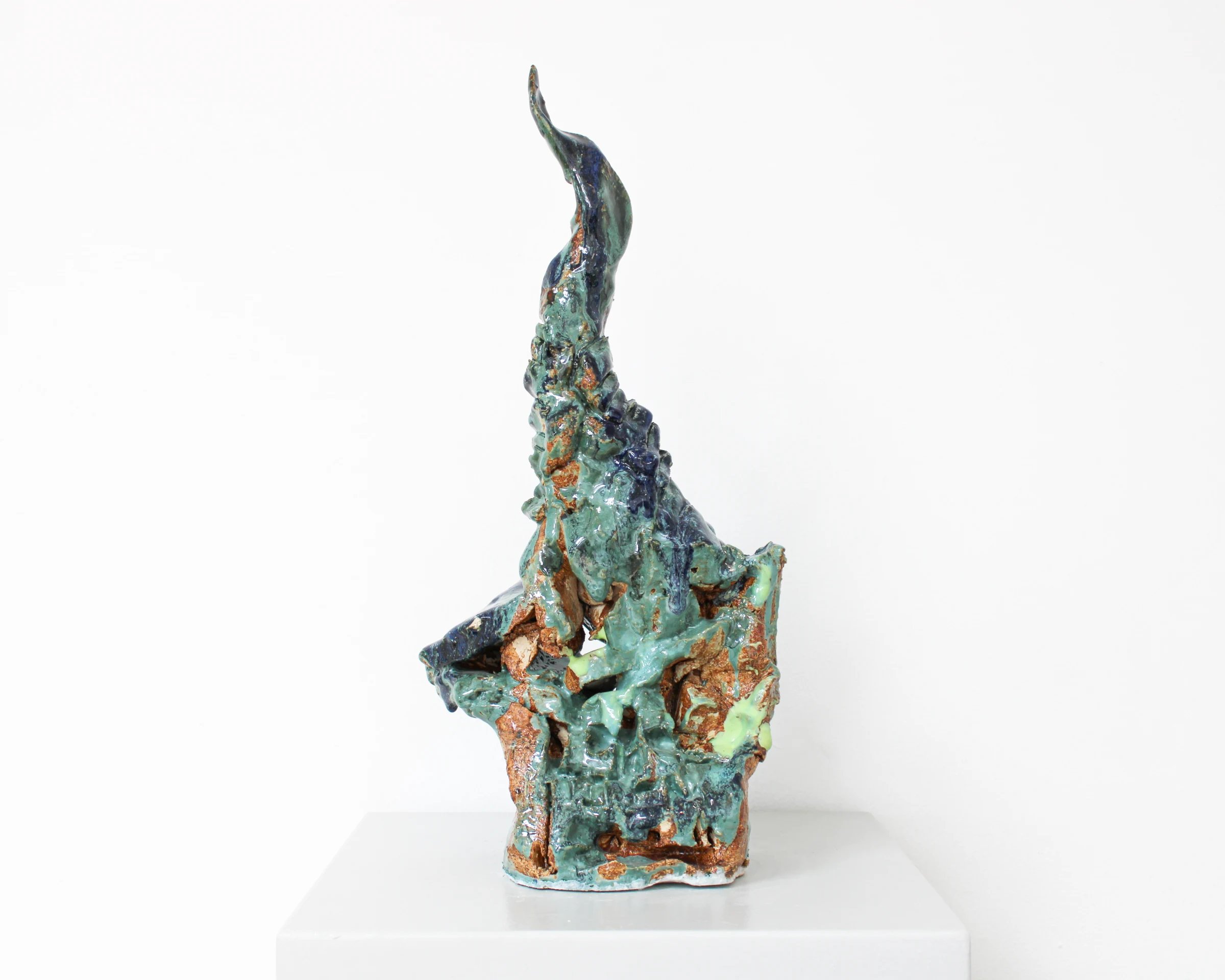 Abstract ceramic sculpture with a tall, twisted, pointed form and textured, layered surface in blue, green, orange, and brown hues, displayed on a white pedestal against a plain white background. Frederik Næblerød exhibition at Alice Folker Gallery, 