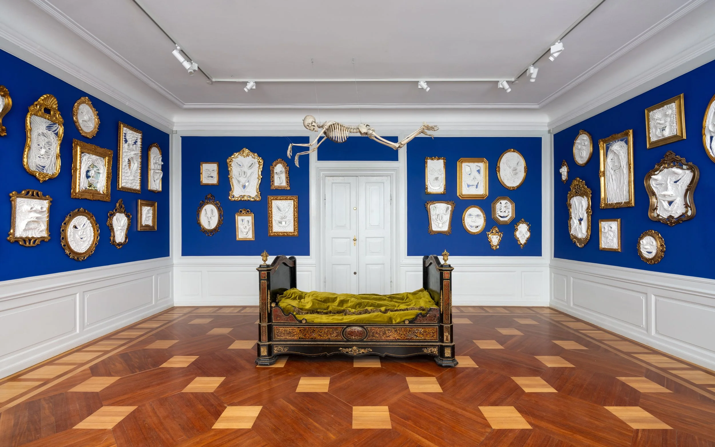 An art gallery featuring blue walls decorated with an array of framed sculptures and masks, and a skeleton hanging from the ceiling with a bed in the center. Frederik Næblerød exhibition at Gl. Holtegaard, Denmark.