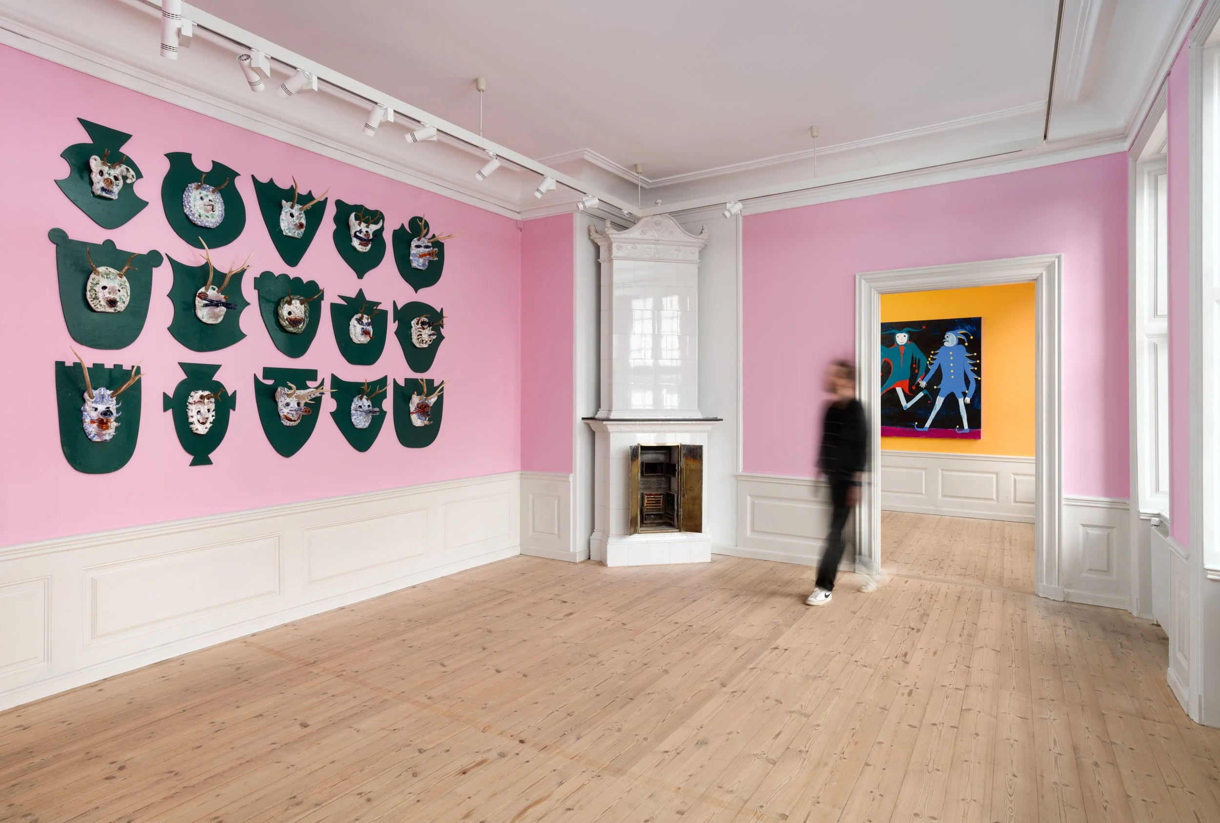 An art gallery with a pink wall displaying mounted decorative masks with antlers, a white fireplace, and a doorway leading to a room with a colorful abstract painting. Frederik Næblerød exhibition at Gl. Holtegaard, Denmark.