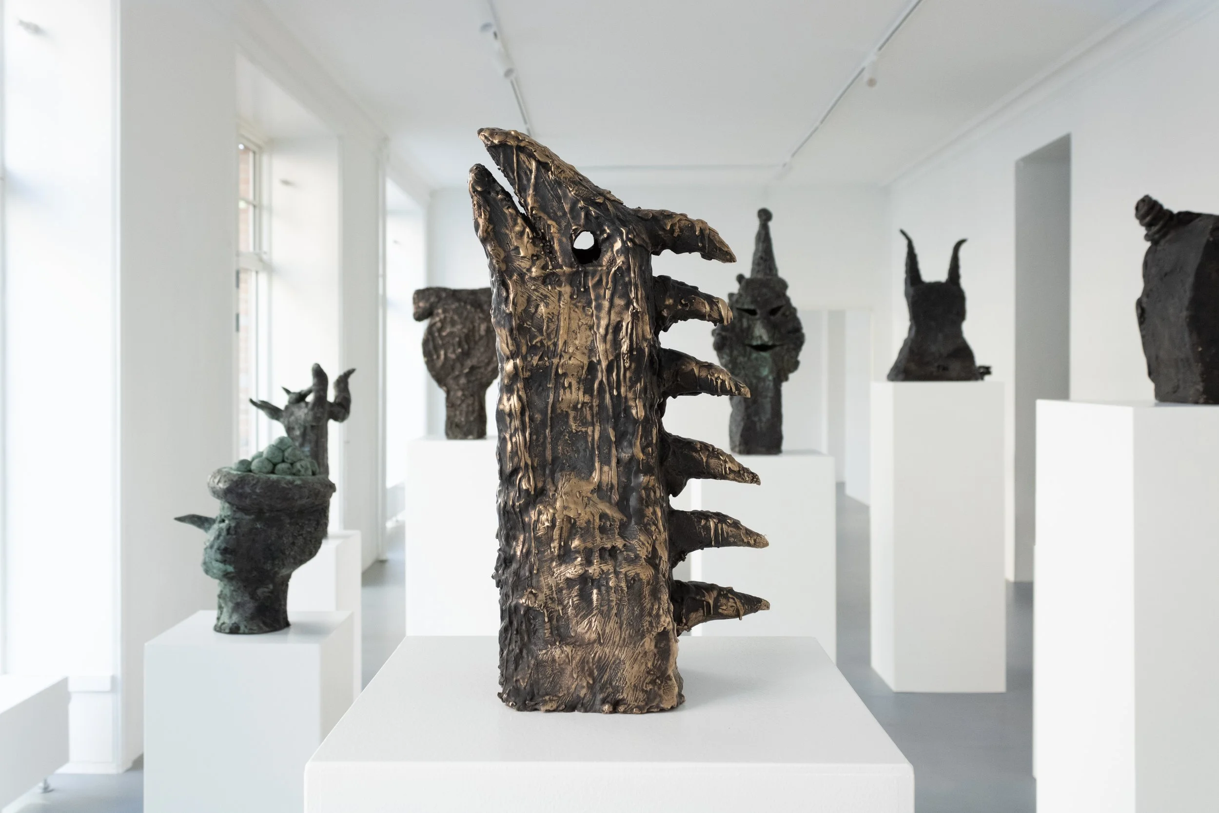 Art exhibition featuring bronze sculptures of fantastical and mythical headpieces on white pedestals in a bright gallery. Frederik Næblerød exhibition at Alice Folker Gallery. 