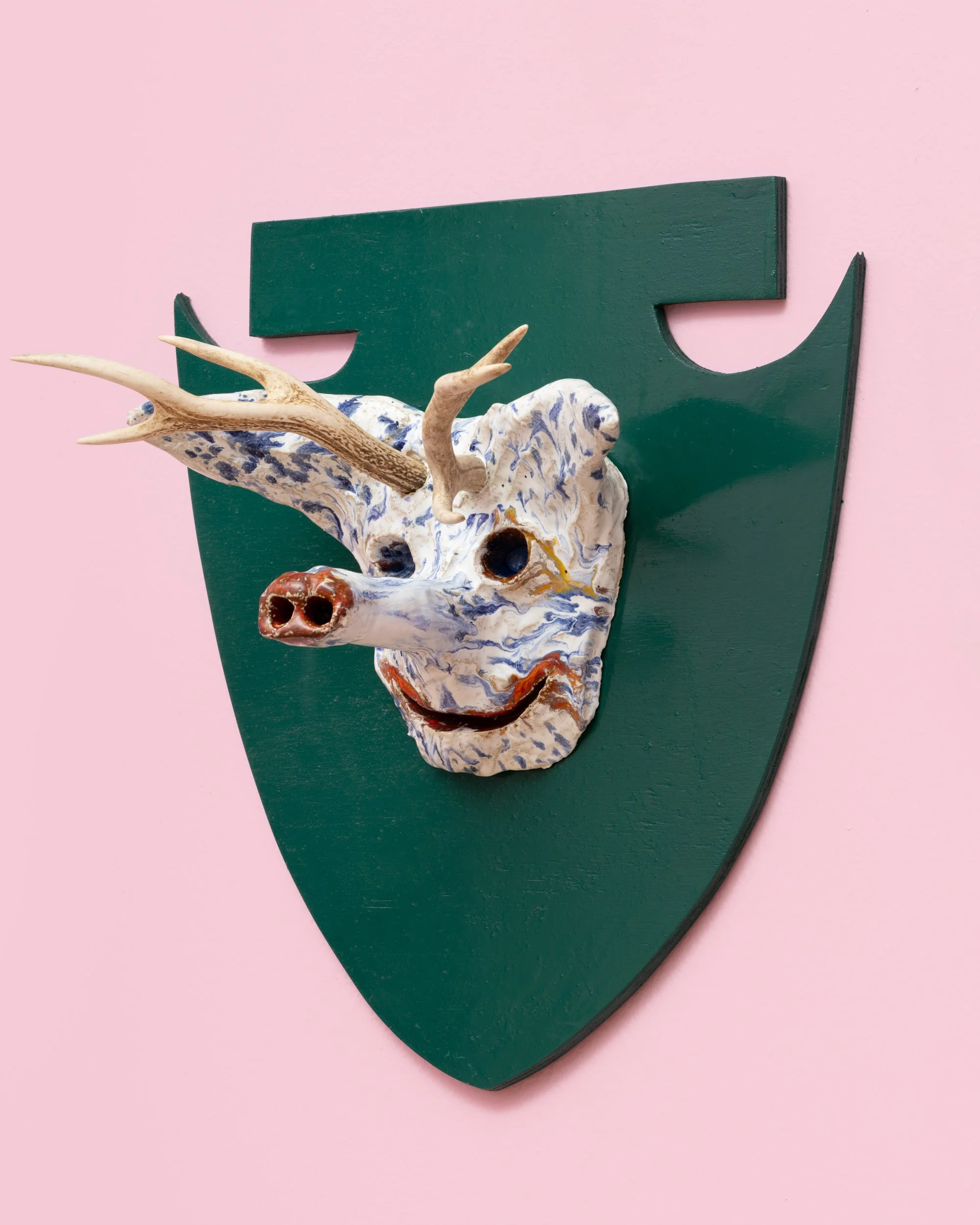 Decorative mounted deer head with painted ceramic facial details on green shield-shaped plaque against pink background. Frederik Næblerød exhibition at Gl. Holtegaard, Denmark.
