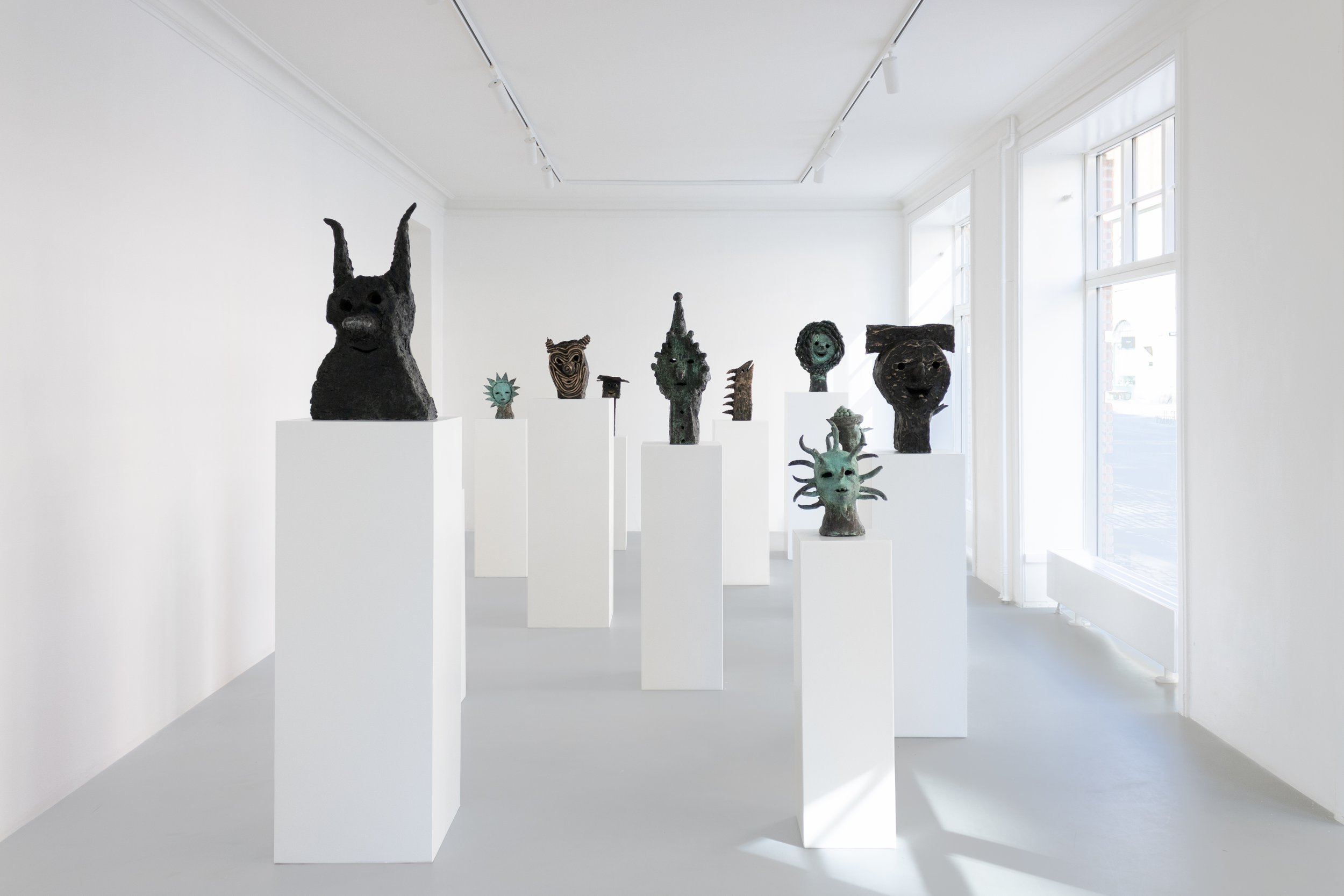 Art exhibition with various dark-colored abstract sculptures on white pedestals in a bright, minimalistic gallery with white walls and large windows. Frederik Næblerød exhibition at Alice Folker Gallery. 