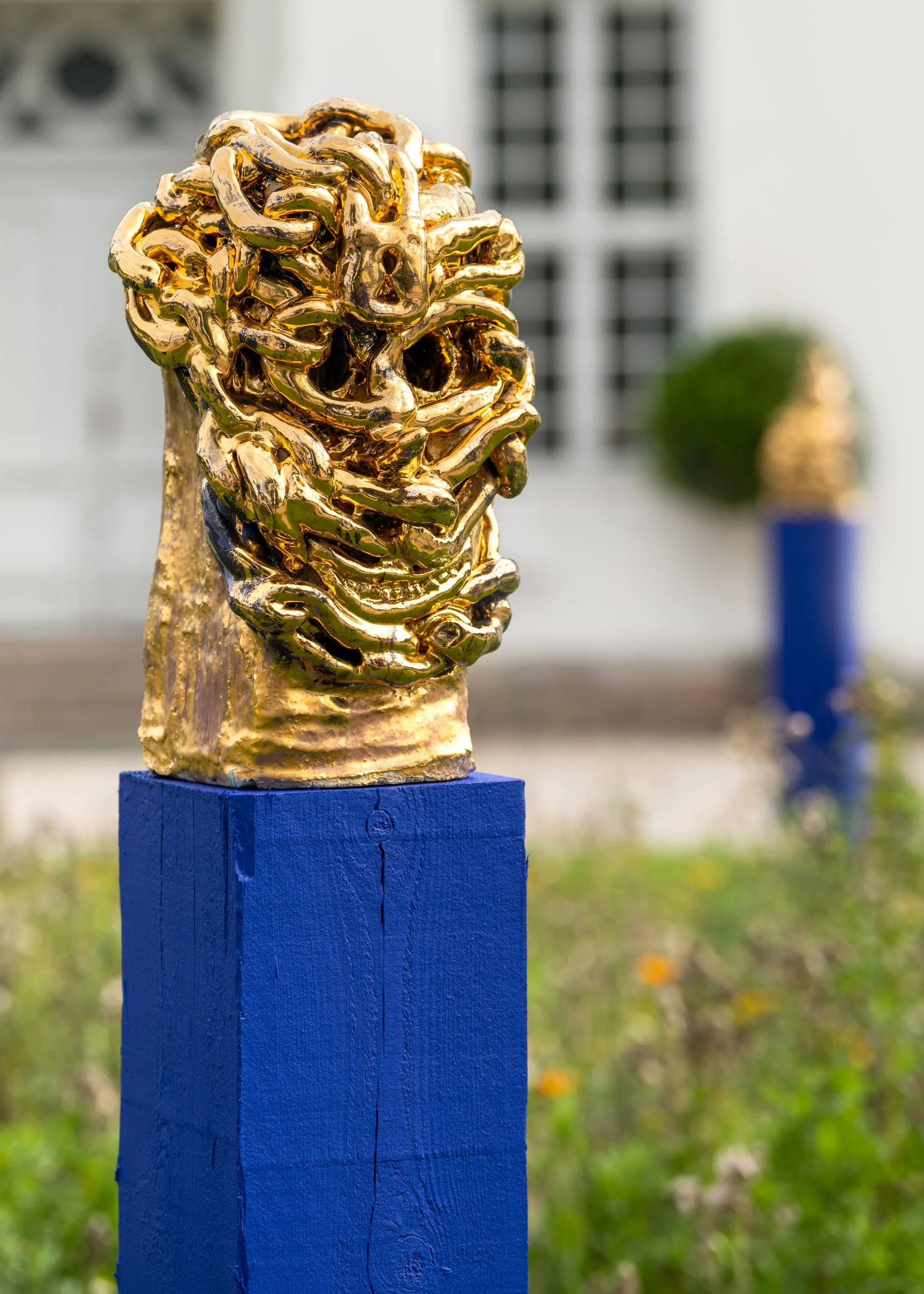 A golden sculpture resembling a lion's head placed on a blue pedestal outdoor, with another sculpture and greenery in the background. Frederik Næblerød exhibition at Gl. Holtegaard, Denmark.
