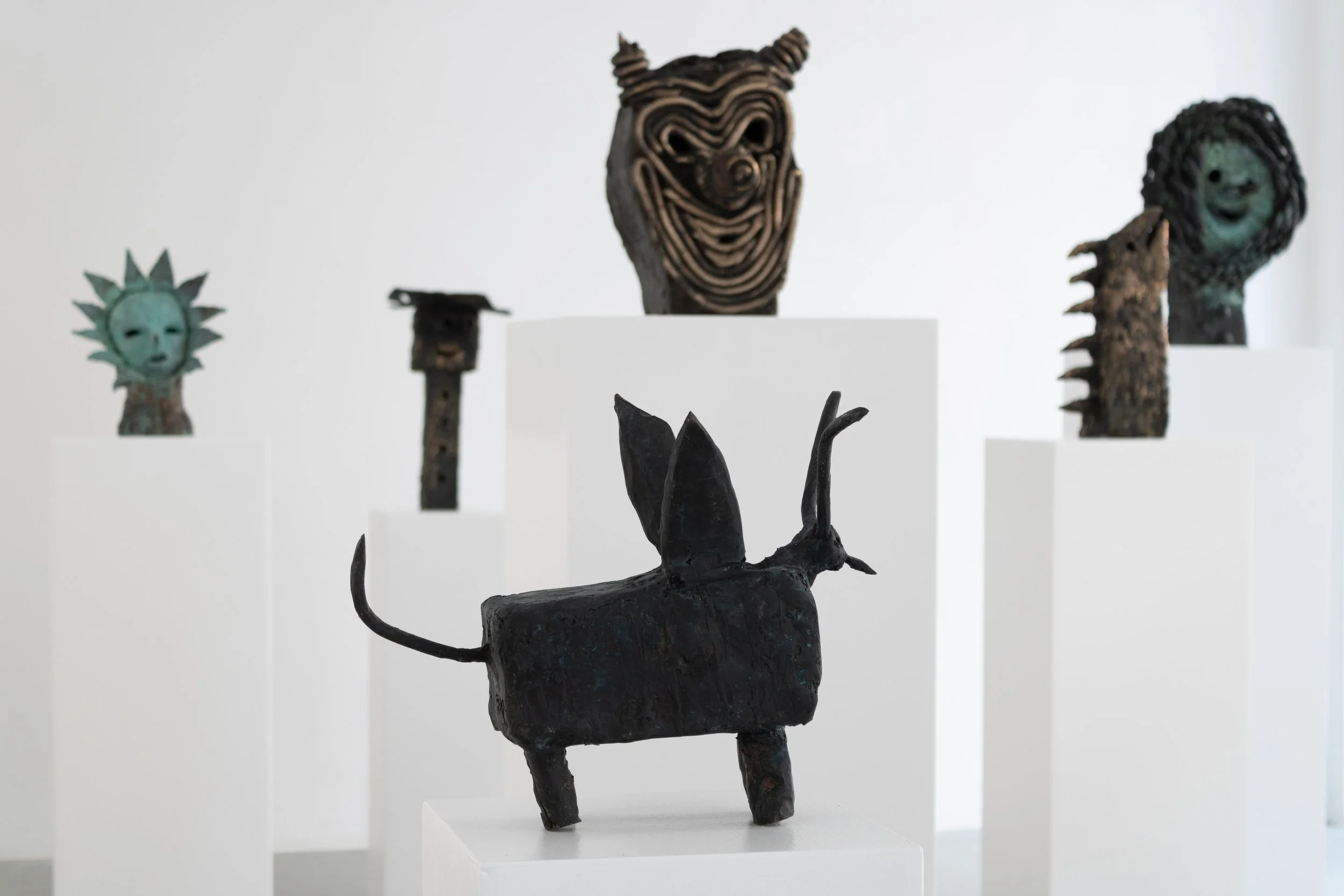 Art sculptures displayed on white pedestals, including a black creature with large ears and a tail in the foreground and various other colorful and textured masks or sculptures in the background. Frederik Næblerød exhibition at Alice Folker Gallery. 