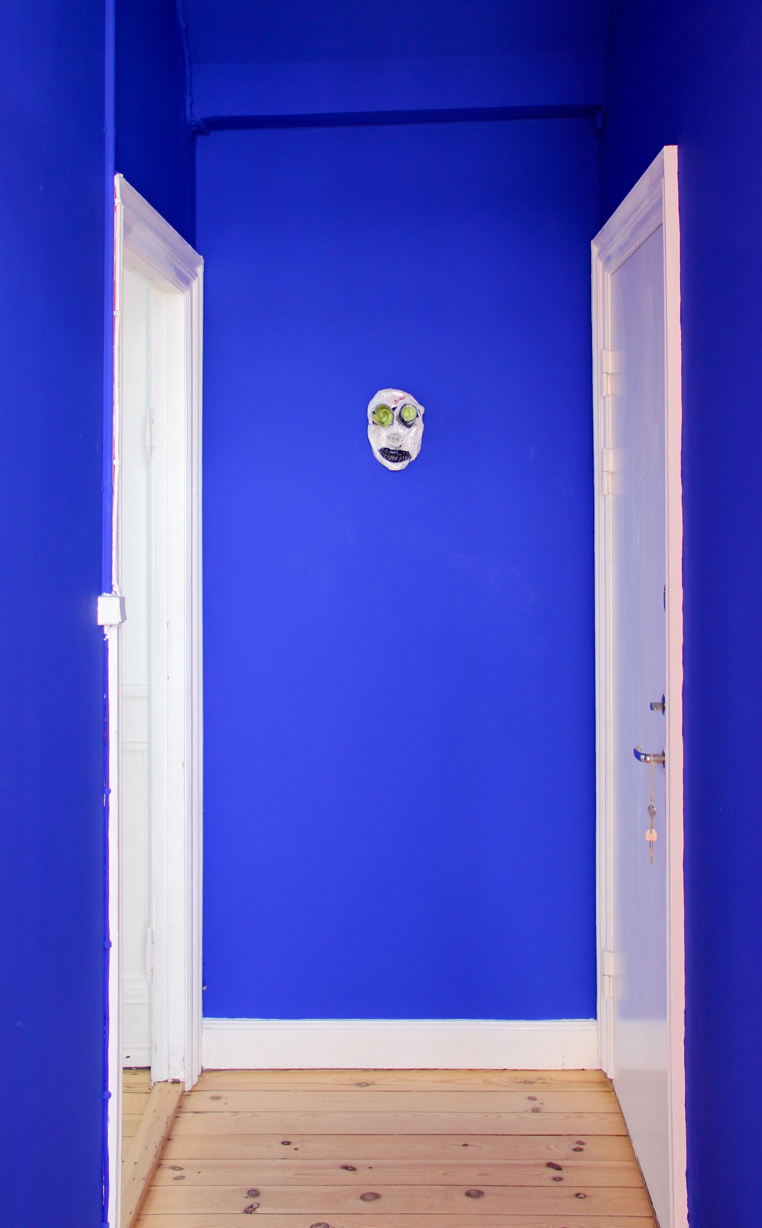 A narrow hallway with bright blue walls, white doorframes, and a light wooden floor. A painted mask with a creepy face is mounted on the wall in the center. Frederik Næblerød exhibition at Alice Folker Gallery, Denmark.