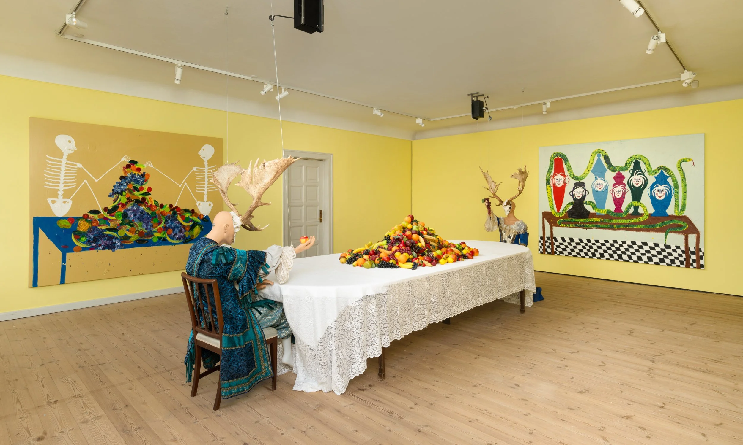 An art installation with mannequins, colorful fruit piled on a table, and paintings on yellow walls. Frederik Næblerød exhibition at Gl. Holtegaard, Denmark.