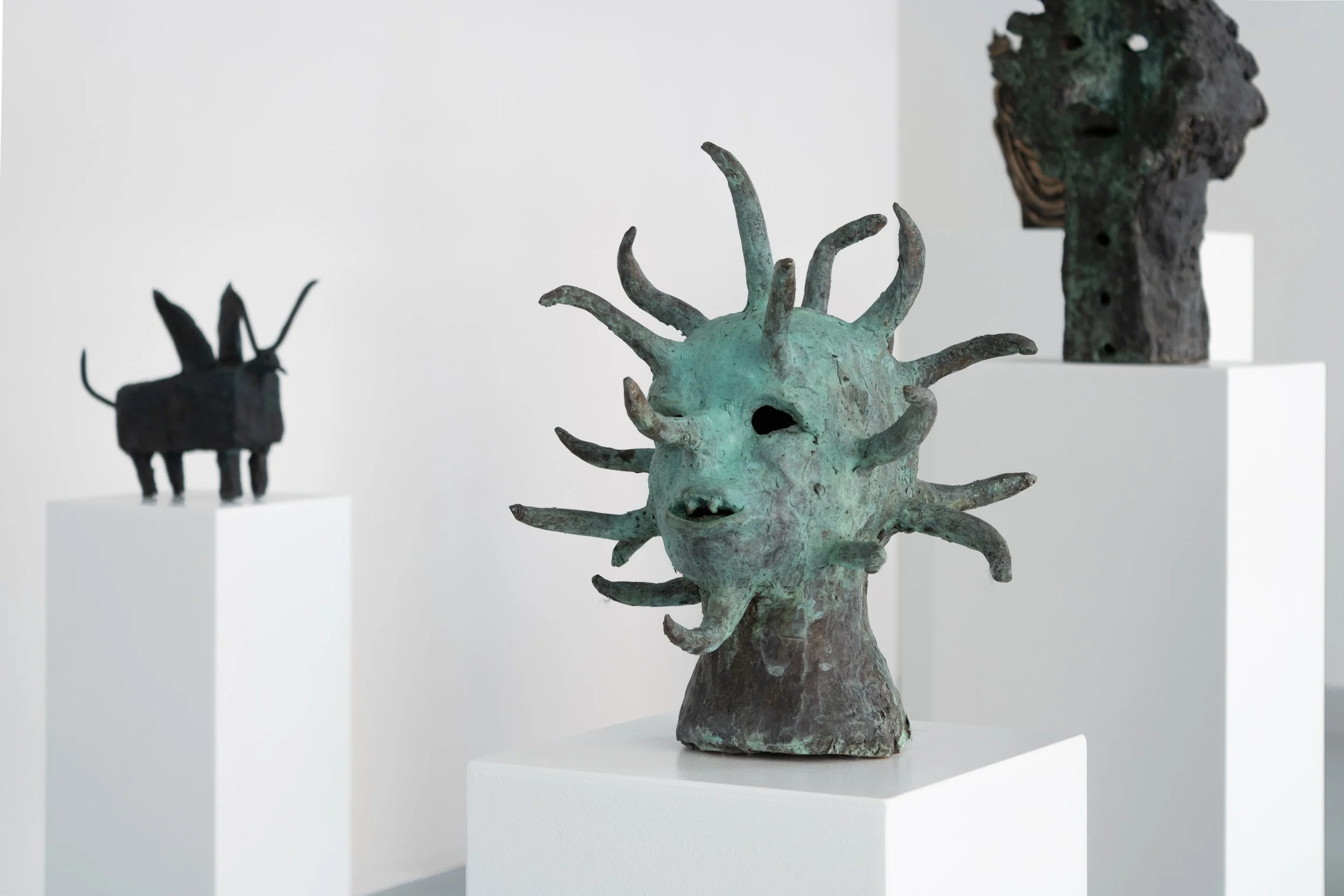 Three abstract sculptures displayed on white pedestals in an art gallery, including a greenish creature with multiple tentacle-like protrusions, a black animal-like figure with long ears and a tail. Frederik Næblerød exhibition at Alice Folker Galler