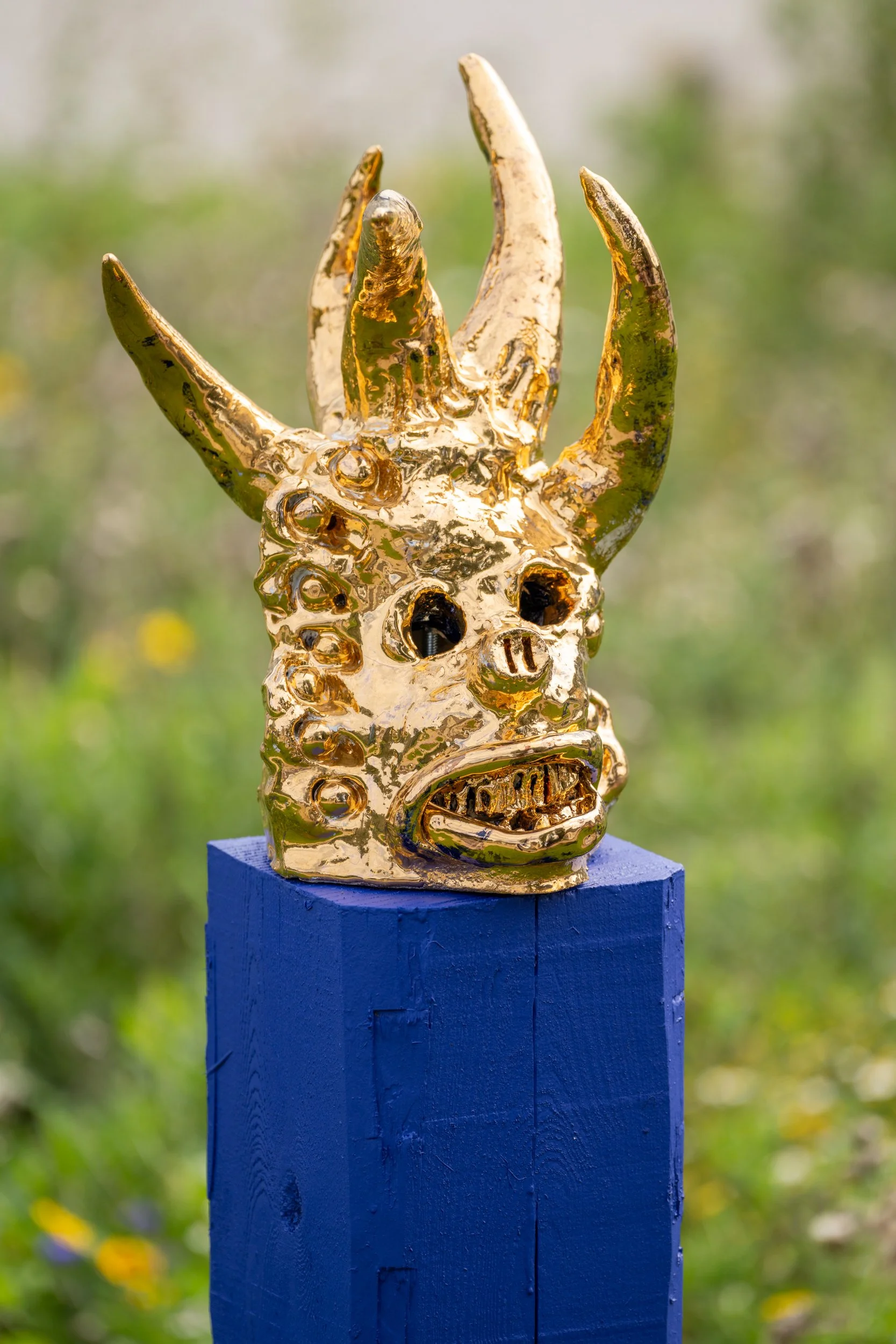 A shiny, gold-colored sculpture of a mythological creature with multiple horns and a face with a furrowed brow, resting on a blue pedestal in an outdoor setting. Frederik Næblerød exhibition at Gl. Holtegaard, Denmark.