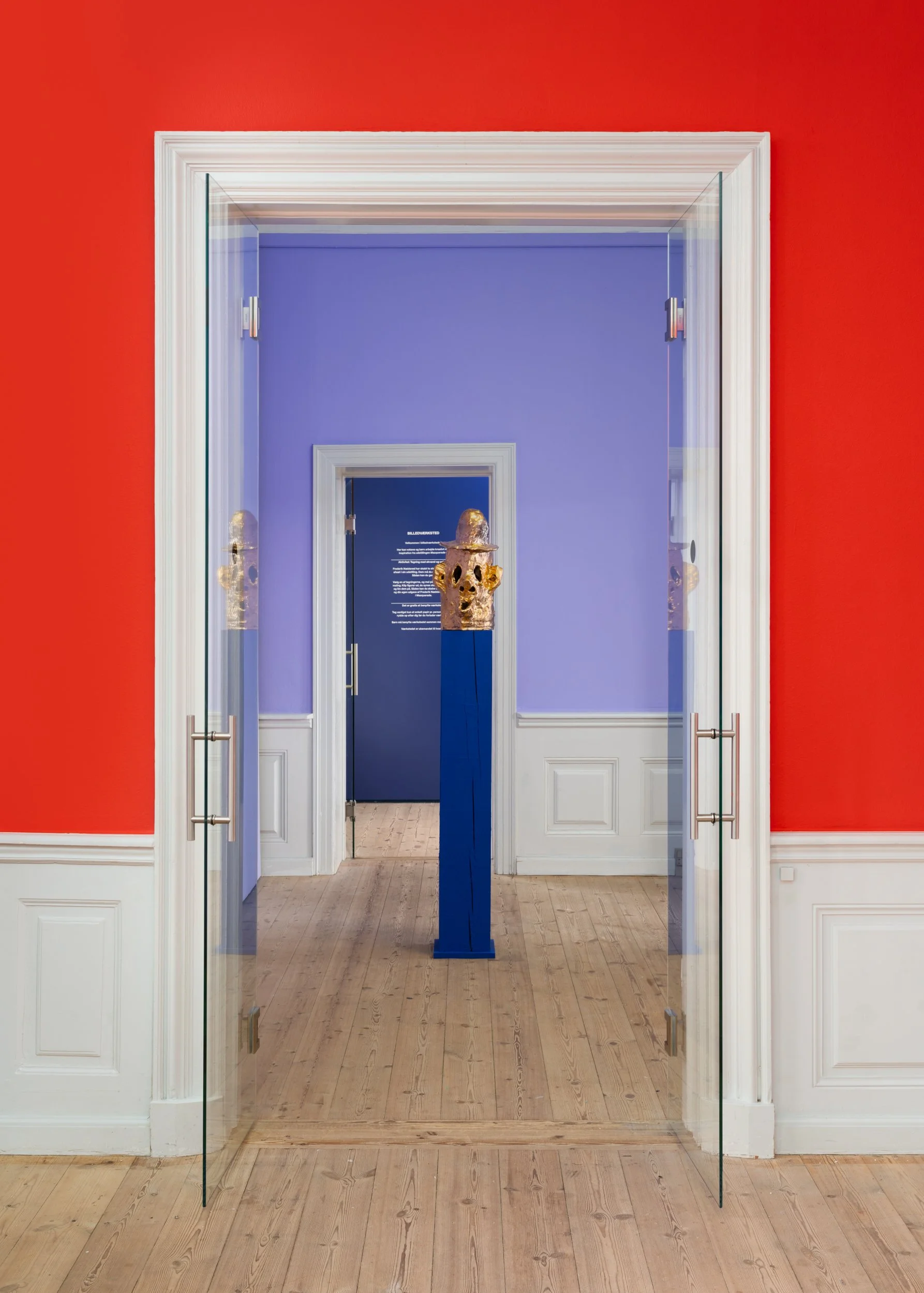View through double glass doors into an art gallery room with purple walls, featuring a sculpture of a gold skull on a blue pedestal. Frederik Næblerød exhibition at Gl. Holtegaard, Denmark.