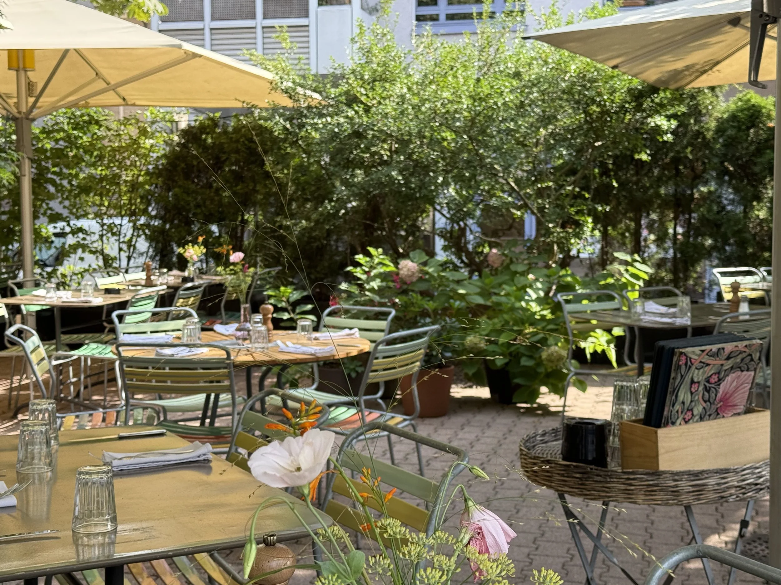 Ein Außenbereich eines Restaurants mit mehreren Tischen und Stühlen, sonnenschirmen, blühenden Pflanzen und grünen Büschen.