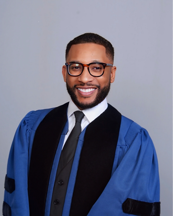 Young man in a graduation cap and gown smiling, wearing glasses against a light gray background.