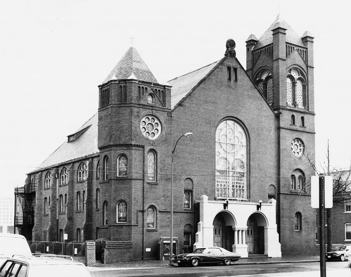 Black and white photo of a large brick church with Gothic architectural features, including tall stained glass windows and a steeple. Several cars are parked in front of the church on the street.