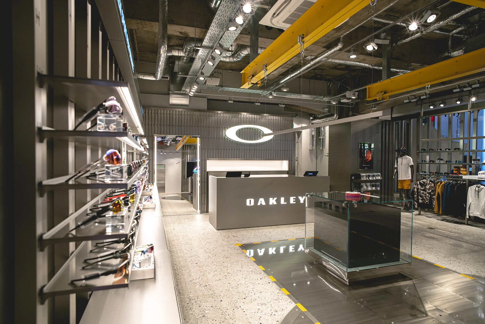 Oakley flagship store Shibuya Japan retail interior wide view showcasing exposed services, yellow gantry beams, eyewear displays, and polished concrete flooring.