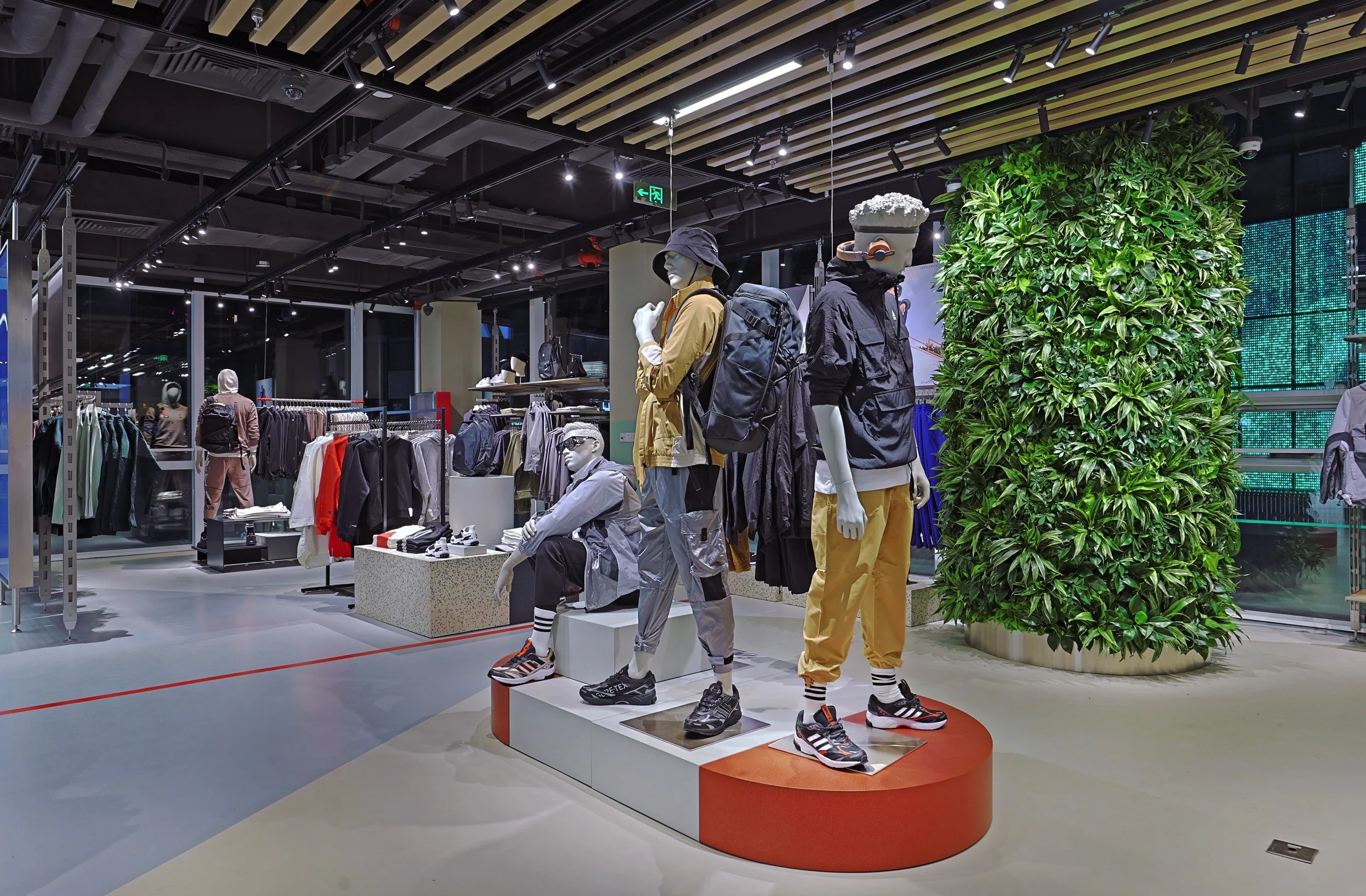 Performance mannequin display inside the Adidas Flagship Sanlitun Beijing featuring layered technical apparel, backpacks, and footwear with a vertical green wall backdrop.