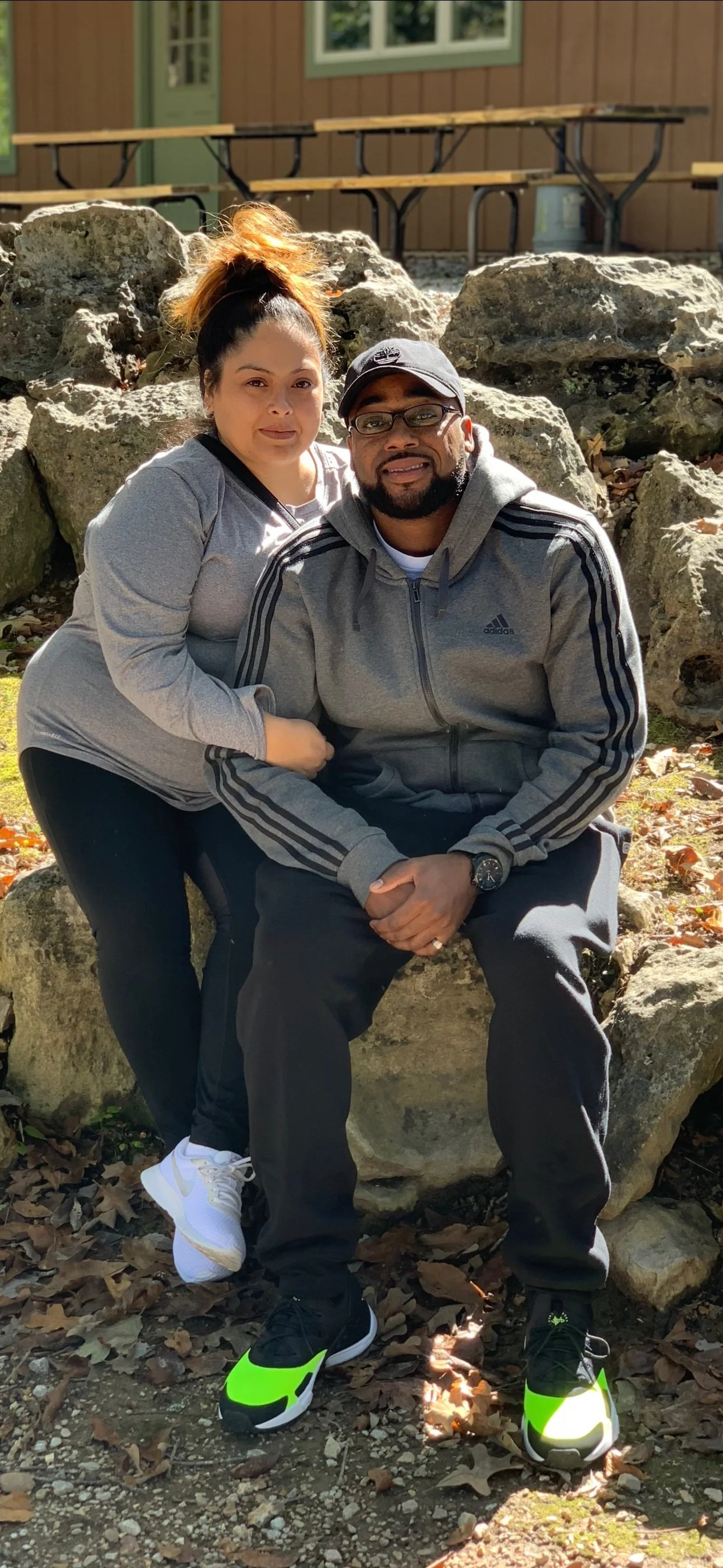 A woman and a man posing together outdoors, sitting on a large rock with fallen leaves around them, in front of a building with a brown exterior and a picnic table in the background.