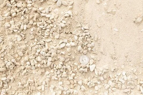 Close-up of crushed light-colored gravel mixed with fine sand, with a coin for scale.