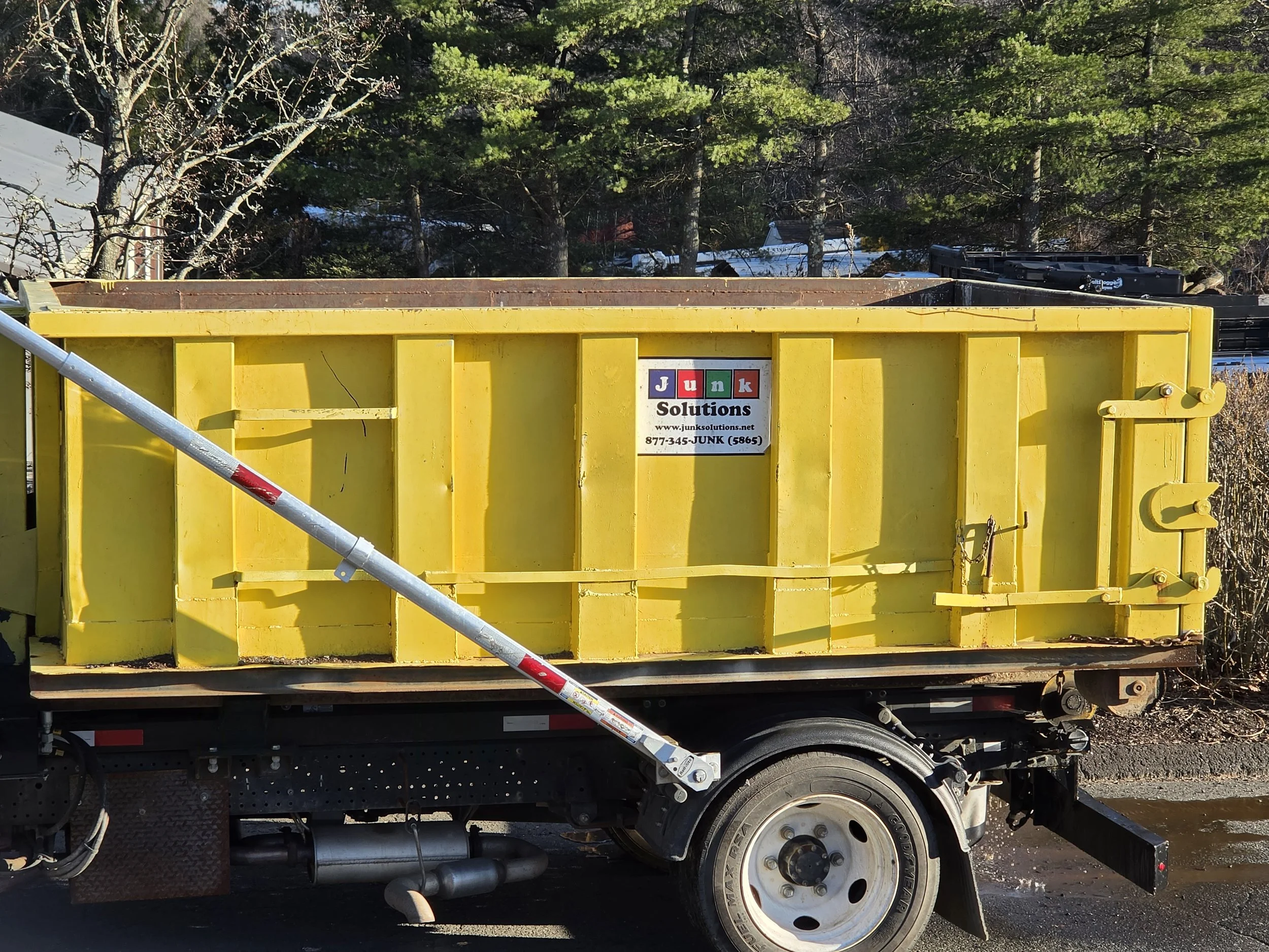 Yellow dumpster truck with a large open top, parked outdoors in a wooded area, with a sign for Junk Solutions attached to the side.