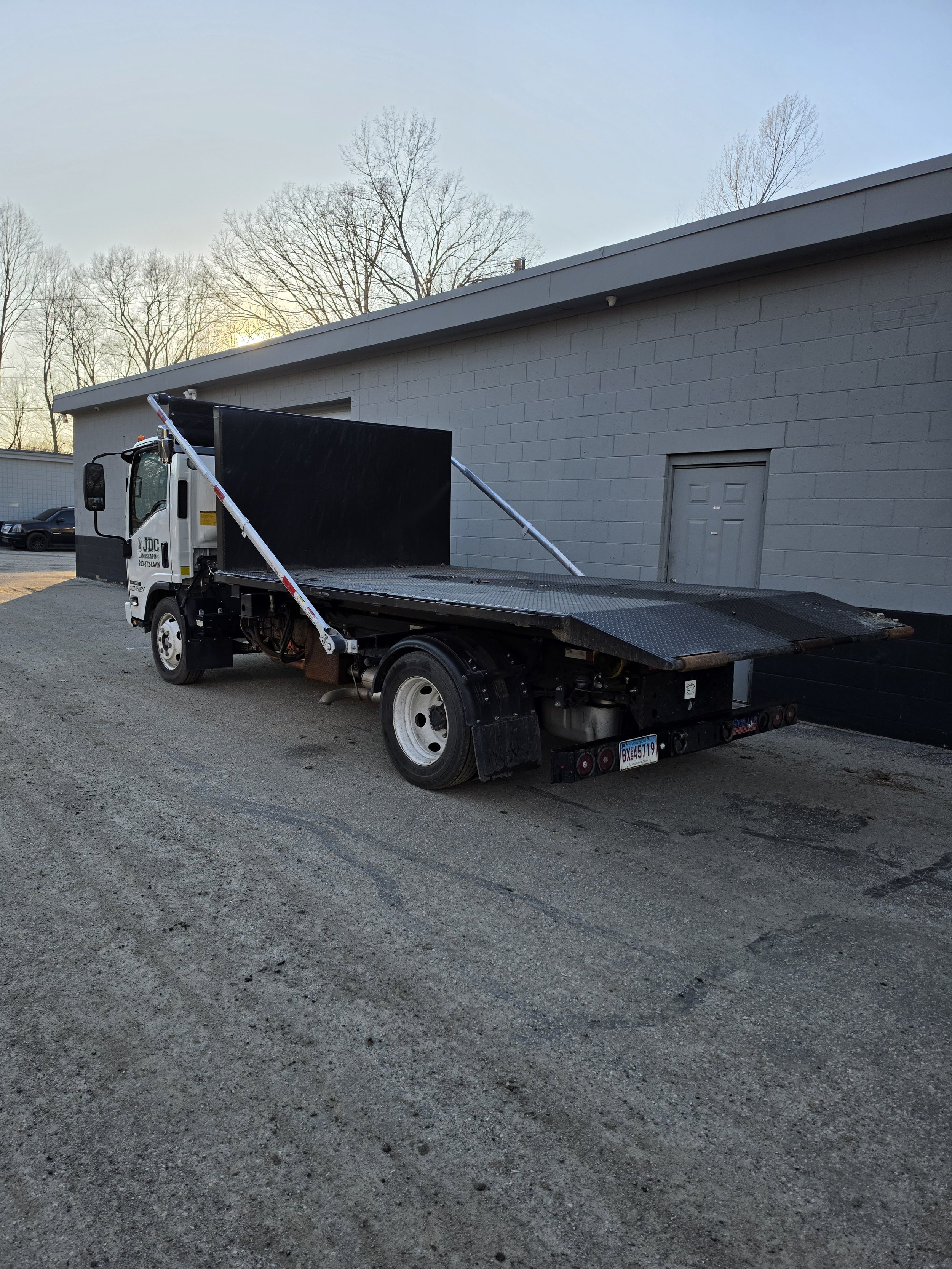A flatbed tow truck parked on gravel against a gray building with a small door. The sky is clear with some trees in the background.