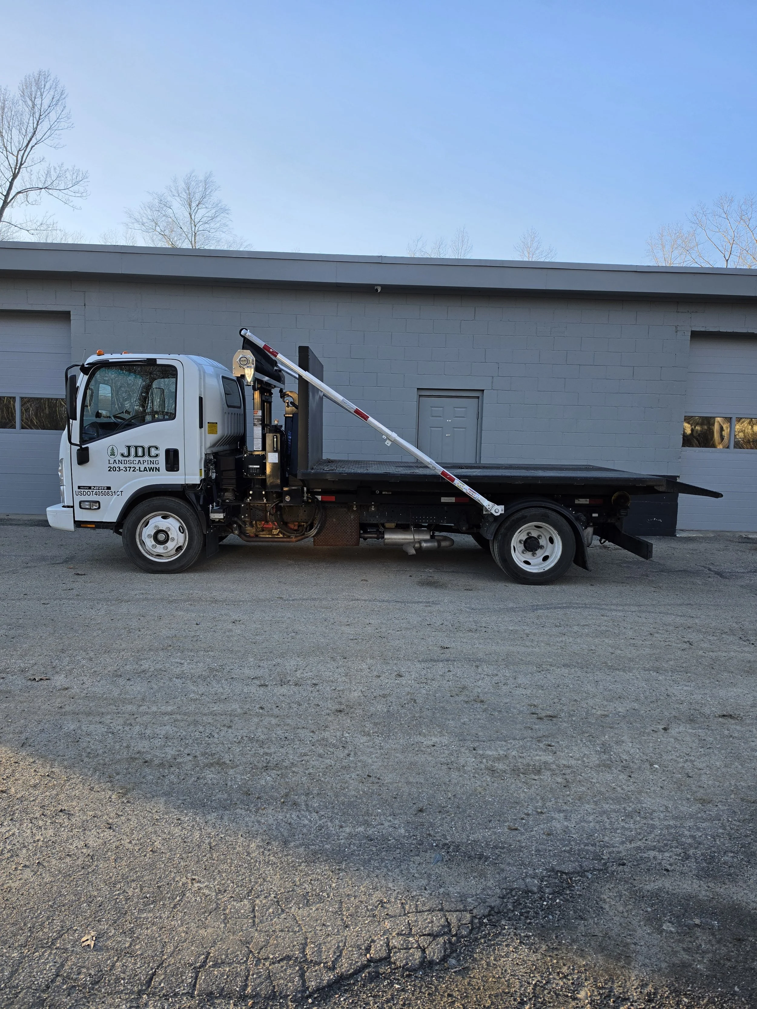 White flatbed truck with JDC Landscaping branding parked on a gravel surface in front of a gray building with closed garage doors and a small window. The truck has a long pole attached to the side.