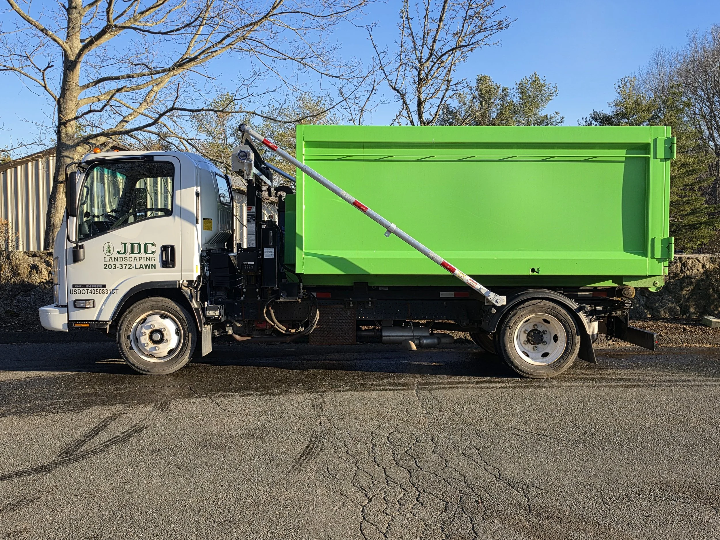 A landscaping truck with a bright green cargo bed parked on a cracked asphalt road, with a tree and sky in the background.