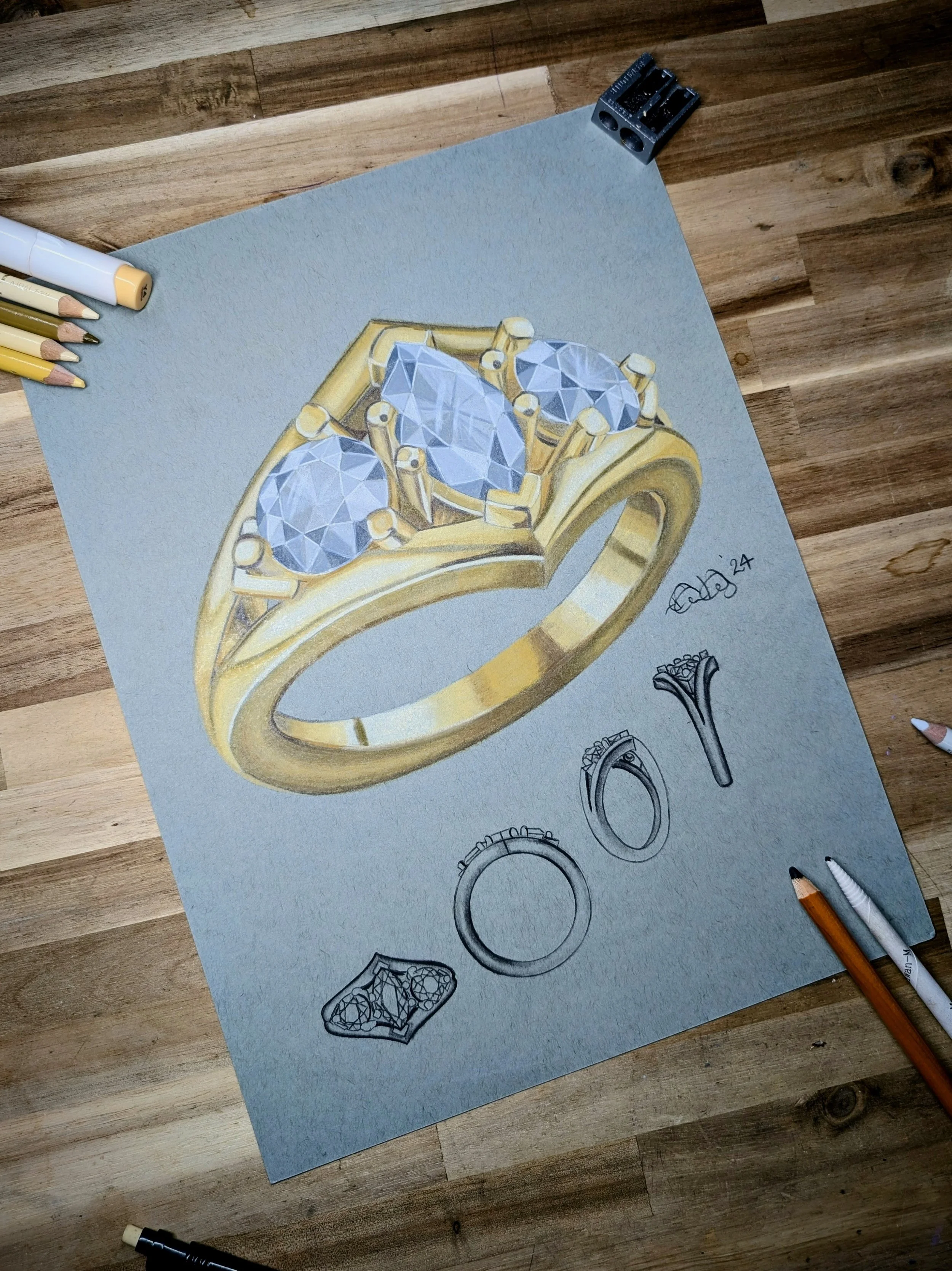 Hand-drawn illustration of a gold ring with three large diamonds on a gray piece of paper. Several smaller sketches of rings are also visible on the paper. The paper is on a wooden surface with colored pencils and pens nearby.