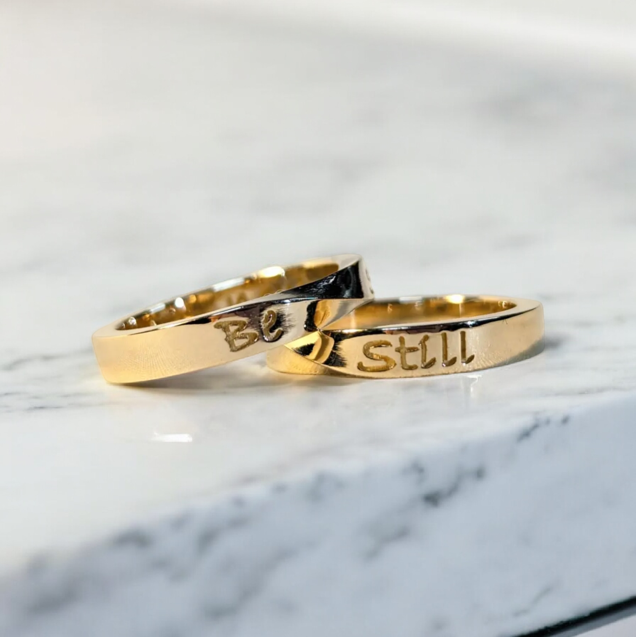 Yellow gold twisted wedding bands with the text "Be Still" on the outside and "& know" on the inside in a carved engraving