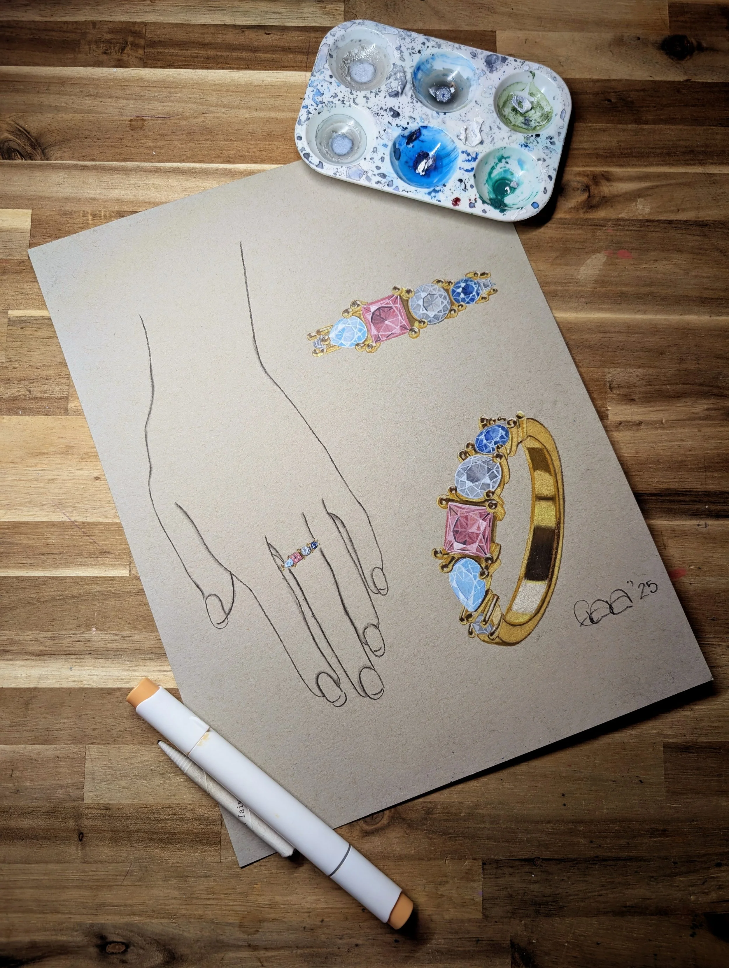 A hand-drawn illustration of a jewelry design on beige paper, featuring a family ring with pink and blue gemstones in gold settings. A palette with paint spots and a white marker are placed on a wooden surface.