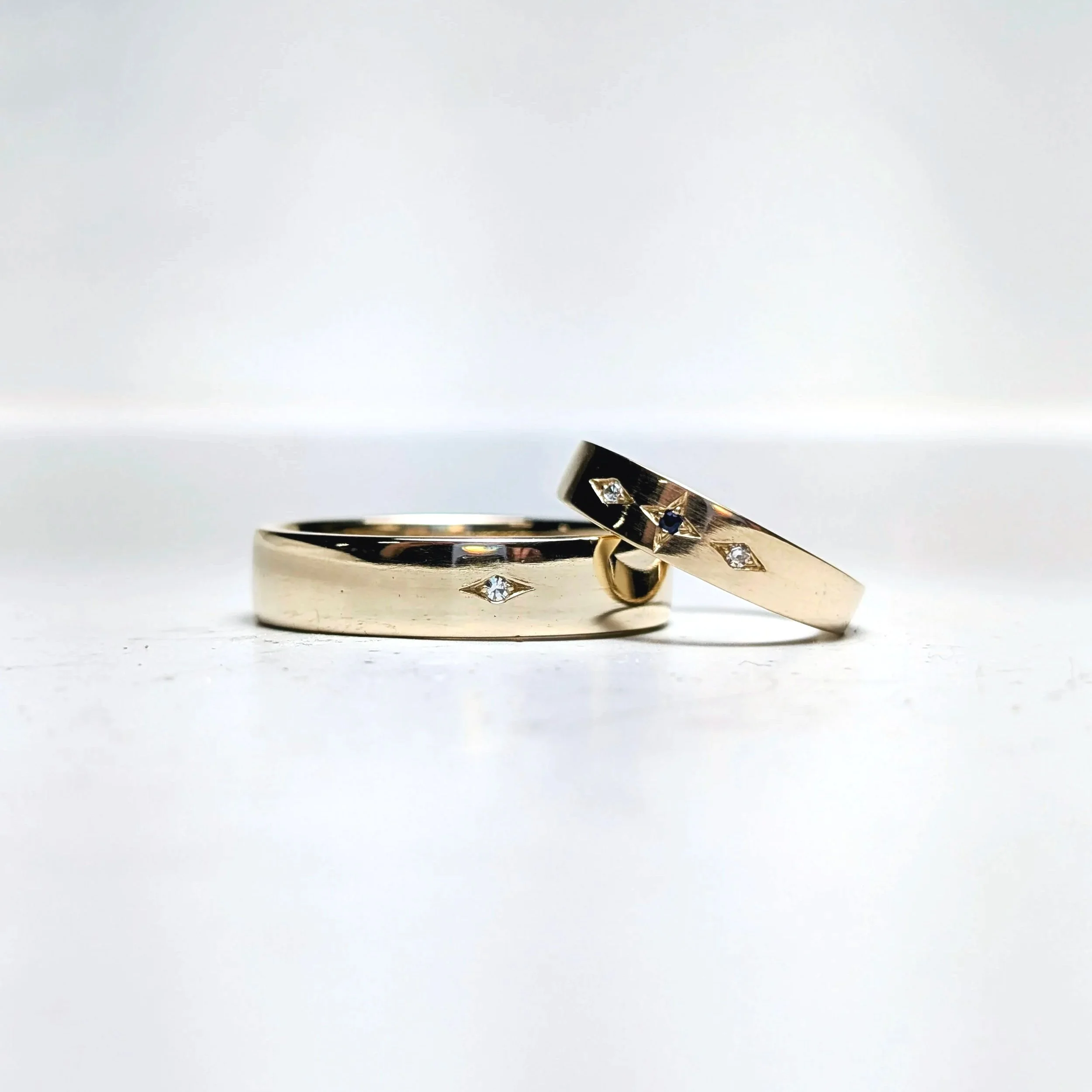 14kt yellow gold wedding bands with a star cut out setting featuring one old mine cut round brilliant diamond on the mens band, and two old mine cut round diamonds and one blue sapphire round brilliant cut bead set on the womens band.